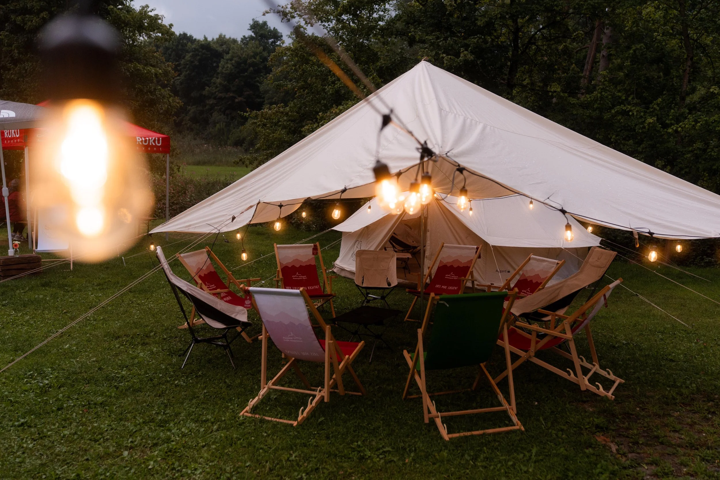 Ein Zelt im Freien bei Sonnenuntergang, umgeben von Campingstühlen und Lichterketten, mit einem Baum im Hintergrund.