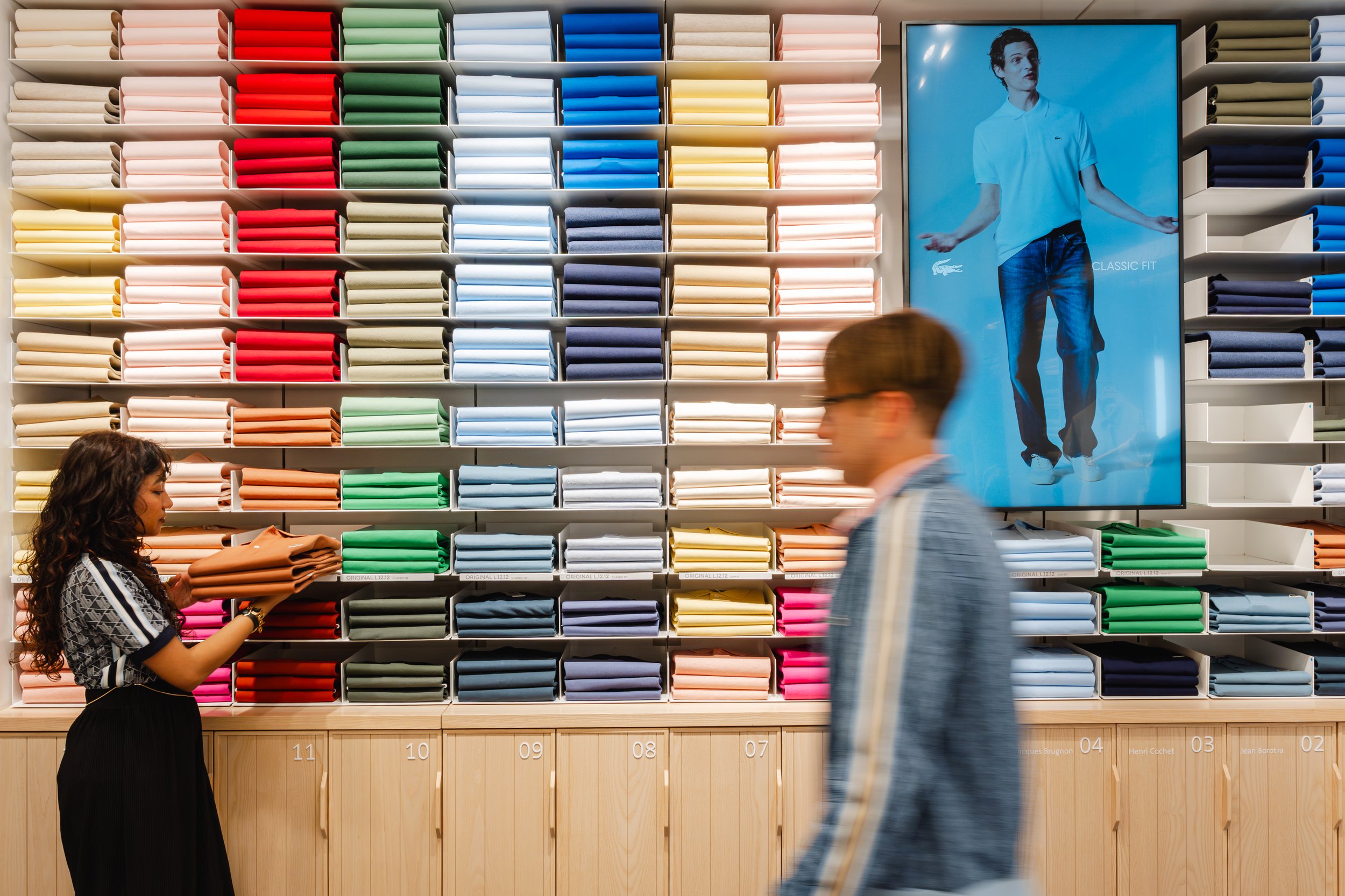 Menschen in einem Stoffladen, eine Frau wählt bunte Stoffe aus, während ein Mann vorbeigeht. Es sind bunte Stoffrollen auf Regalen. Ein großes Bildschirm zeigt eine Person in einem weißen Hemd, die spricht.
