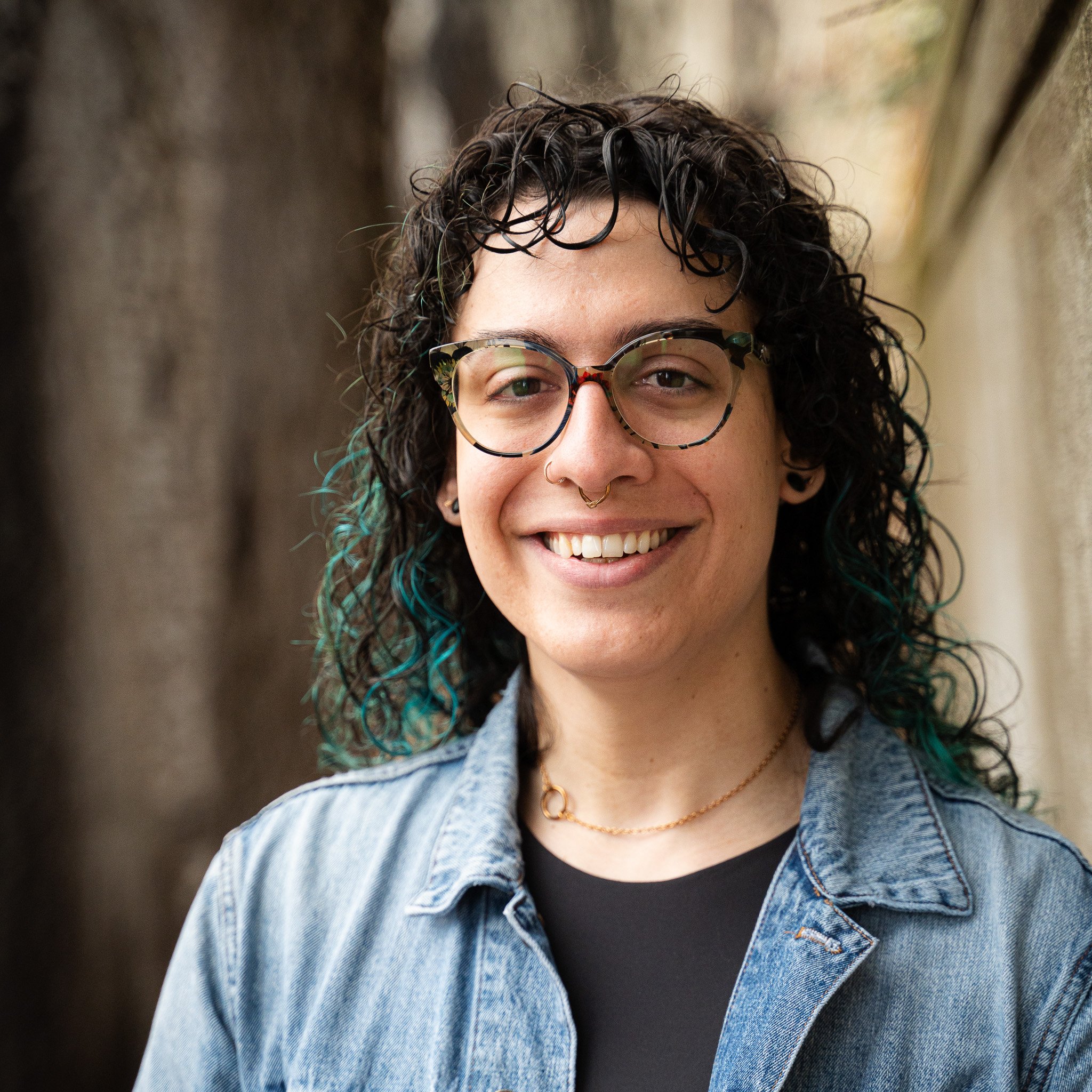 A smiling person with curly hair wearing glasses, a nose ring, a black shirt, a denim jacket, and a gold necklace.