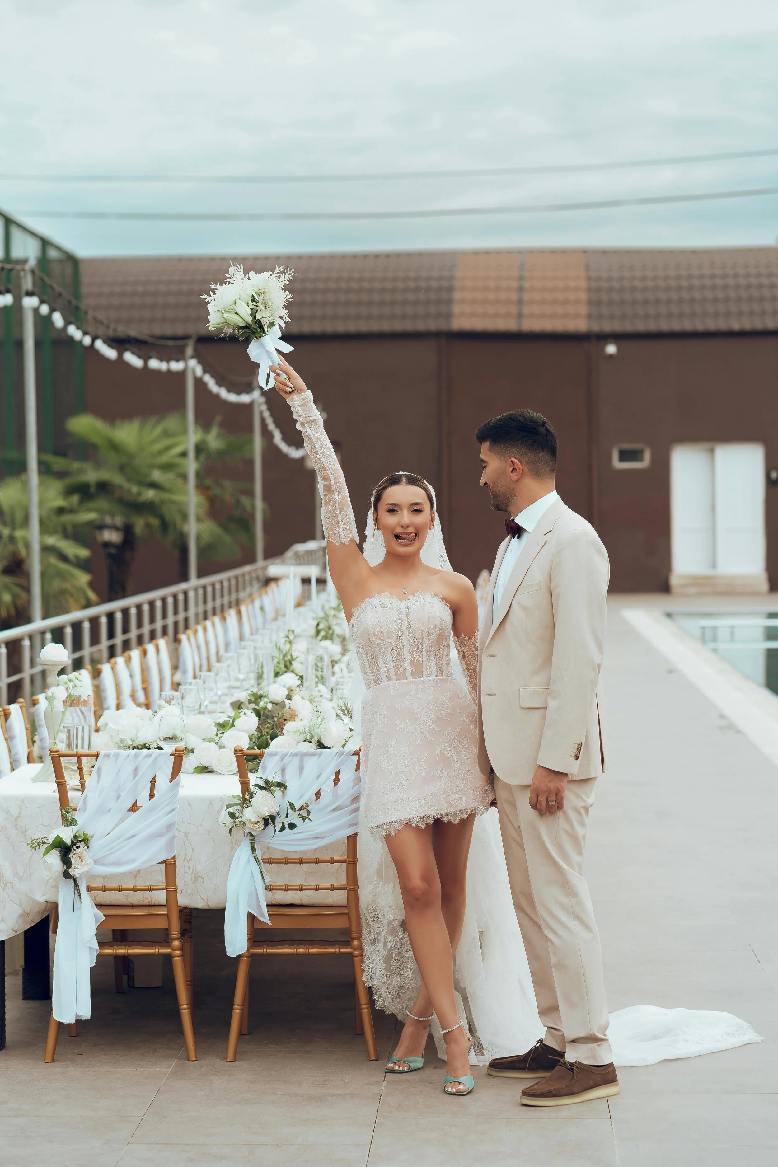 A newlywed couple stands at a decorated outdoor wedding reception. The bride, wearing a lace dress and turquoise heels, holds a bouquet up high and smiles. The groom, in a beige suit with a bow tie, looks at her. The reception area has a long table with white floral arrangements and gold chairs with white fabric accents, set against a backdrop of a brown building, a pool, and string lights under a cloudy sky.