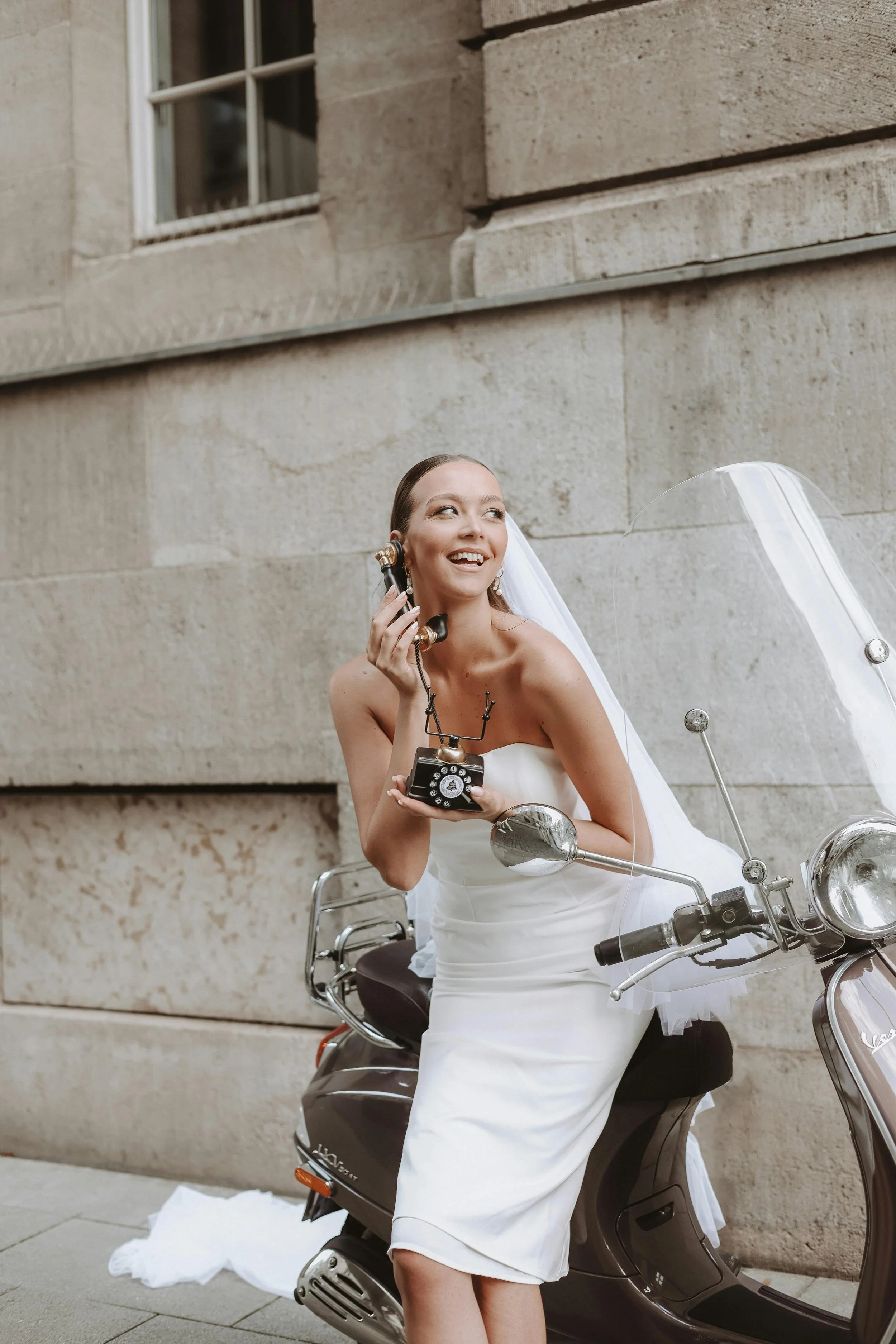 A woman in a white dress sitting on a black scooter, talking on a black rotary phone, with a building wall in the background.