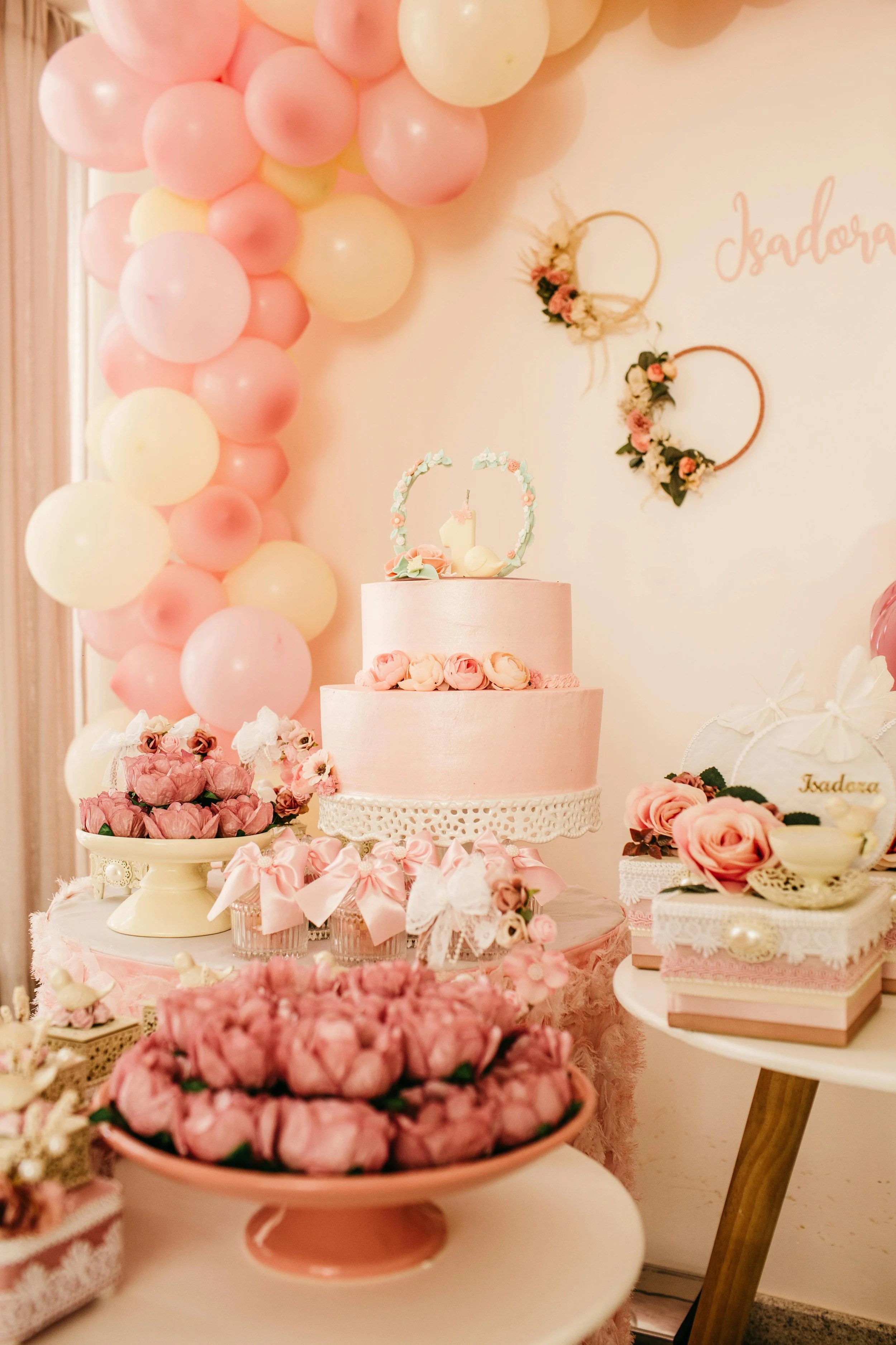 Decorative pink and cream balloons and floral arrangements for a celebration, featuring a pink cake with floral decorations and a backdrop with pink lettering and floral hoops.