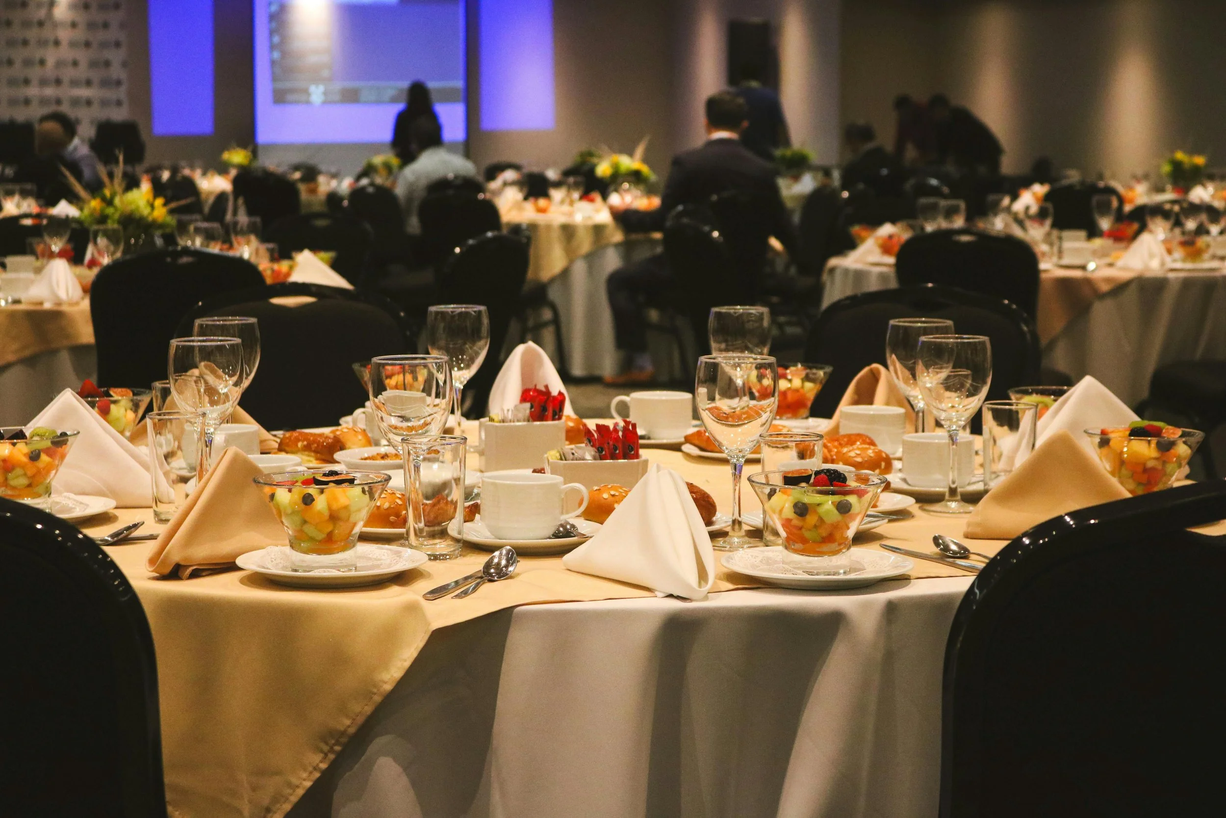 A banquet hall with round tables set for a formal event, decorated with flowers, glassware, and napkins, with a stage and projection screens in the background.