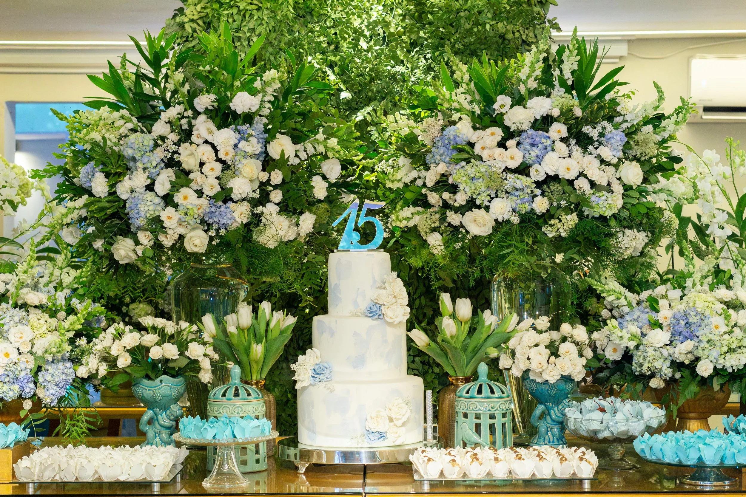 A lavish celebration display with a white cake and an elaborate arrangement of white and blue flowers, greenery, and decorative elements celebrating 15 years.