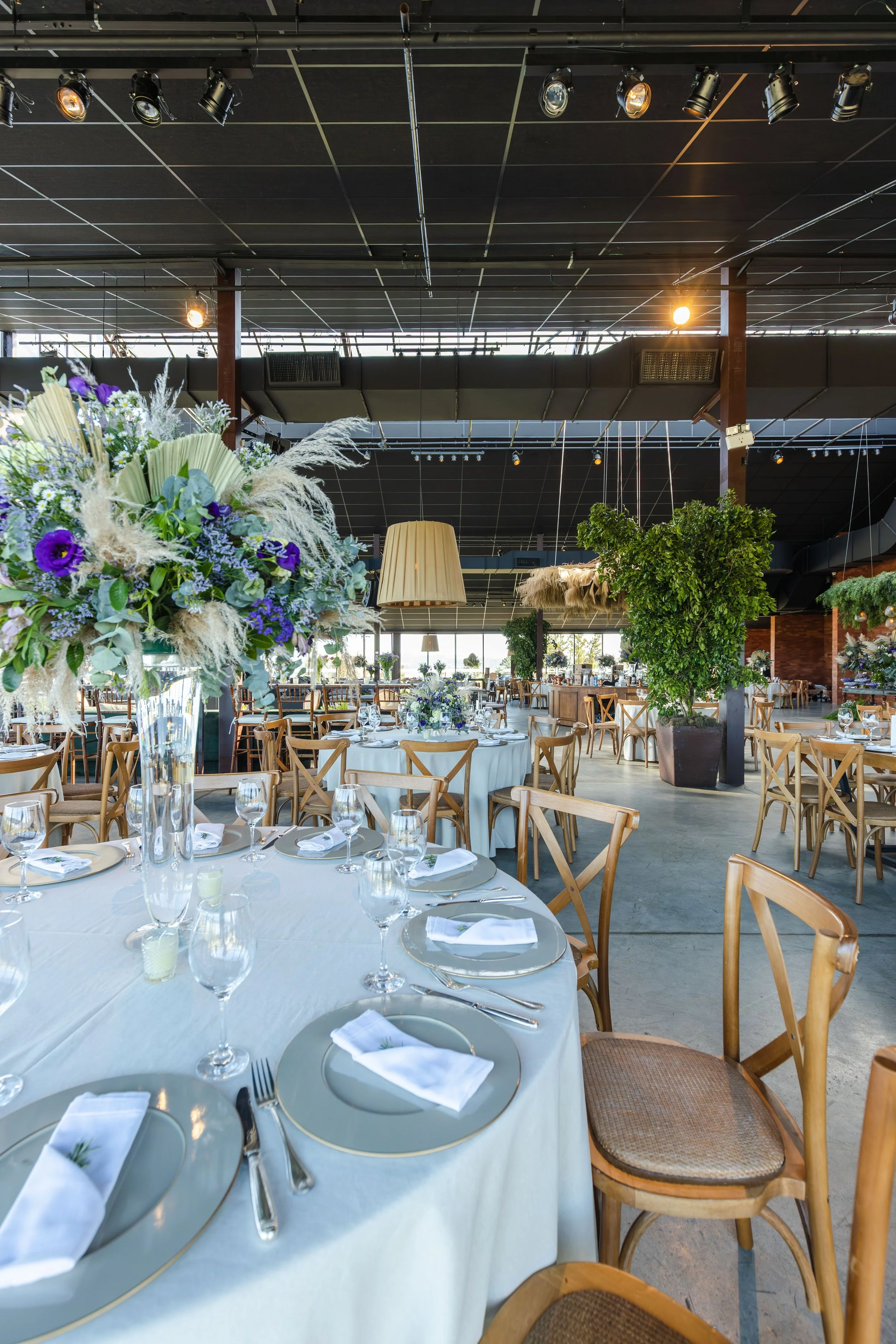 Event space decorated with floral arrangements, tables set with white tablecloths, plates, glasses, and napkins, and wooden chairs surrounding the tables, with a large window letting in natural light and a high ceiling with black panels and stage lights.