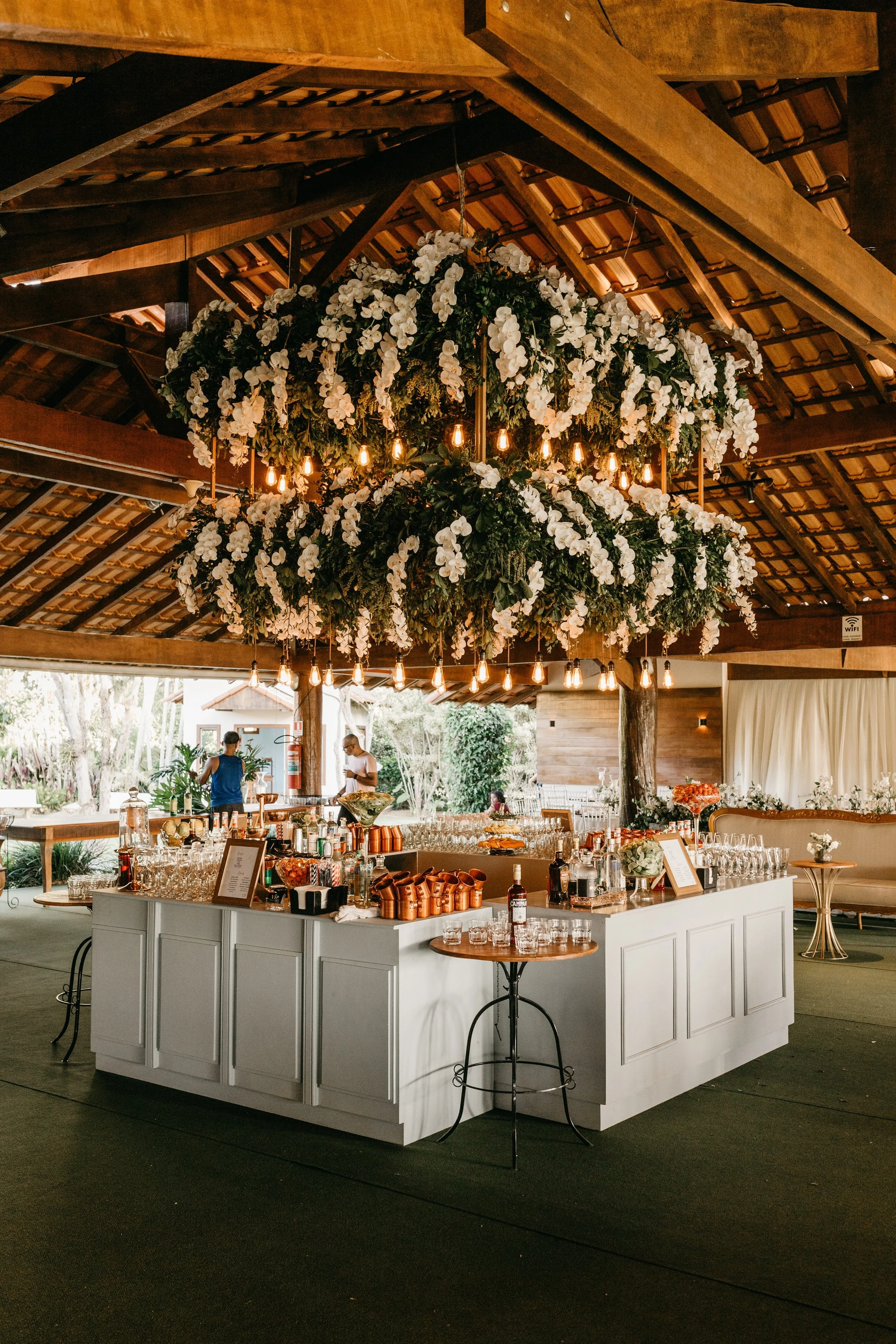 A large indoor event space with a high wooden ceiling featuring exposed beams. A circular bar with white paneling is decorated with glassware, bottles, and coffee cups. Above the bar, there is a grand floral chandelier with white flowers and hanging light bulbs. In the background, two people are preparing drinks, and there is outdoor greenery visible through open walls.