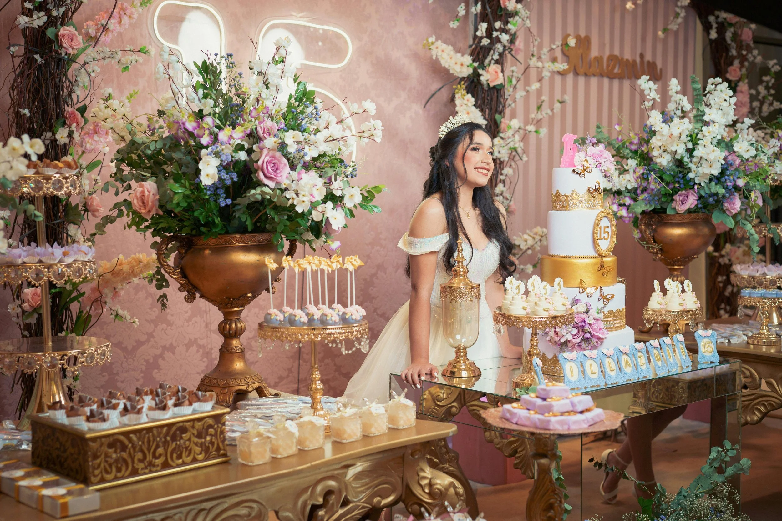 A young woman wearing a tiara and an off-shoulder dress is smiling at a birthday celebration. The event features a table decorated with a three-tiered birthday cake, floral arrangements in gold vases, and various desserts. The background includes pink striped wallpaper and floral decorations.