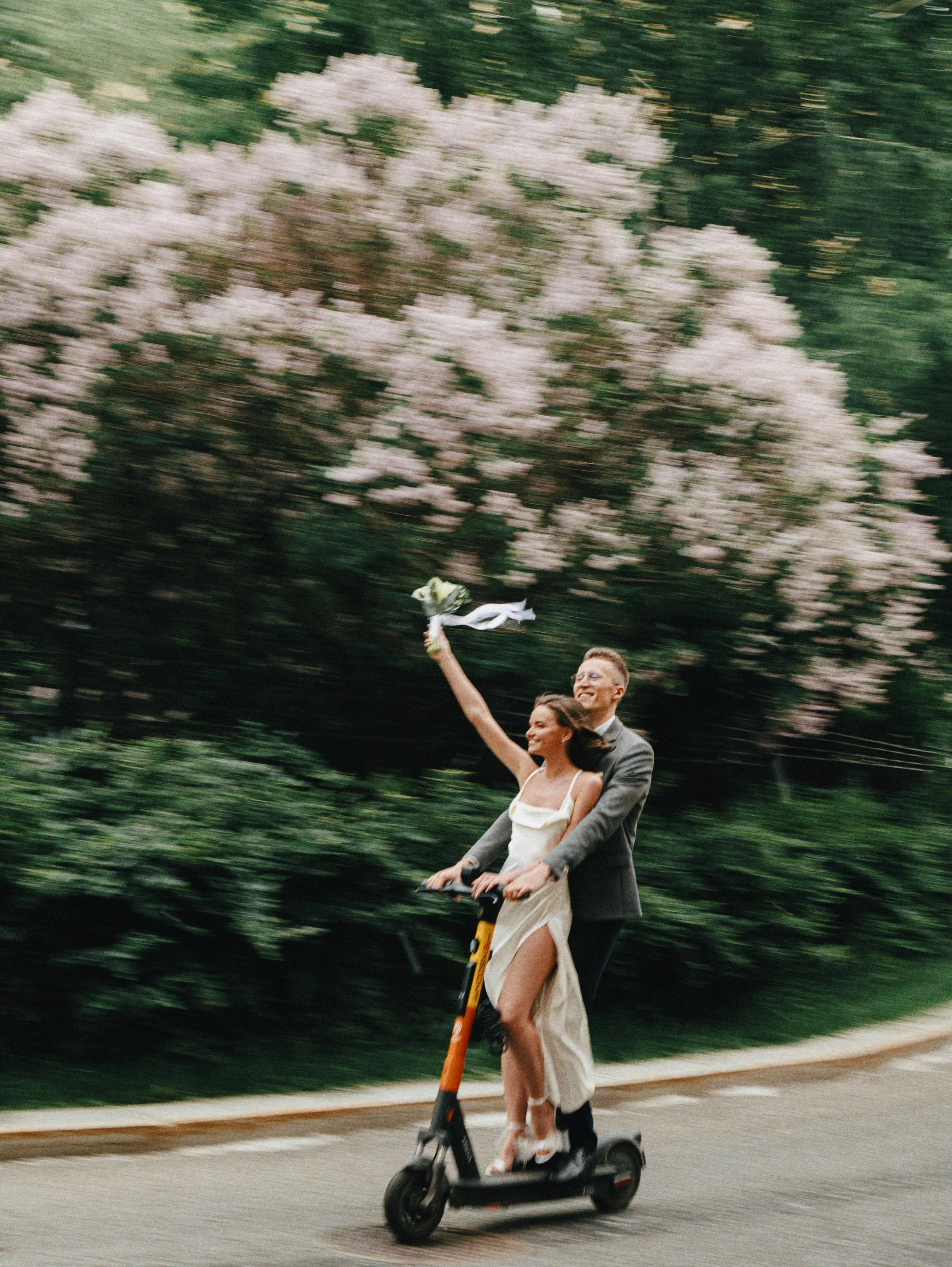 A couple riding an electric scooter on the sidewalk with a lush green hedge and blooming pink and white flowers in the background. The man is dressed in a suit and the woman in a white dress.