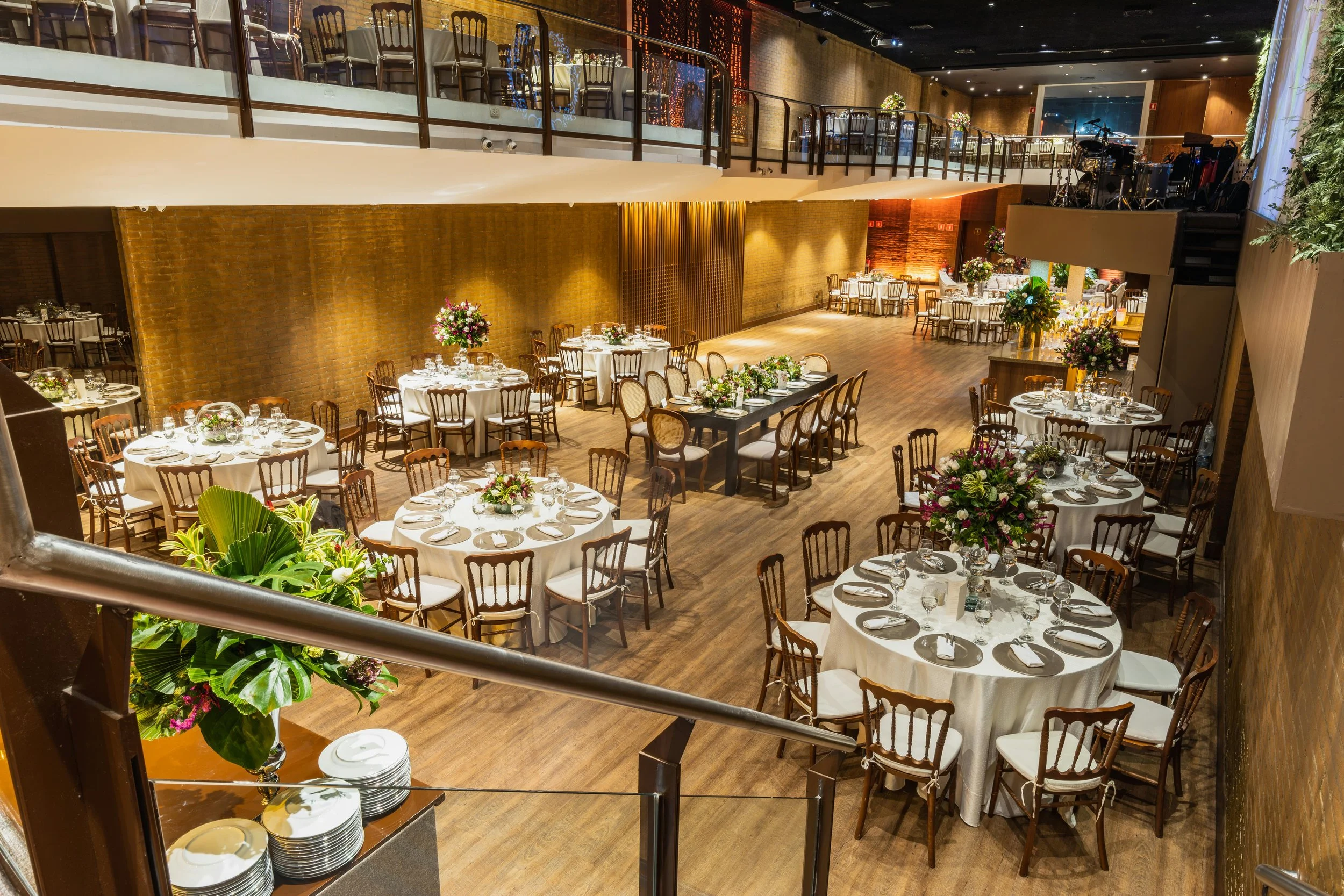A spacious banquet hall decorated for a formal event with round and rectangular tables covered in white tablecloths, floral centerpieces, and place settings. The hall has an upper balcony with chairs, brick walls, wooden flooring, and a stage area with musical instruments and equipment.