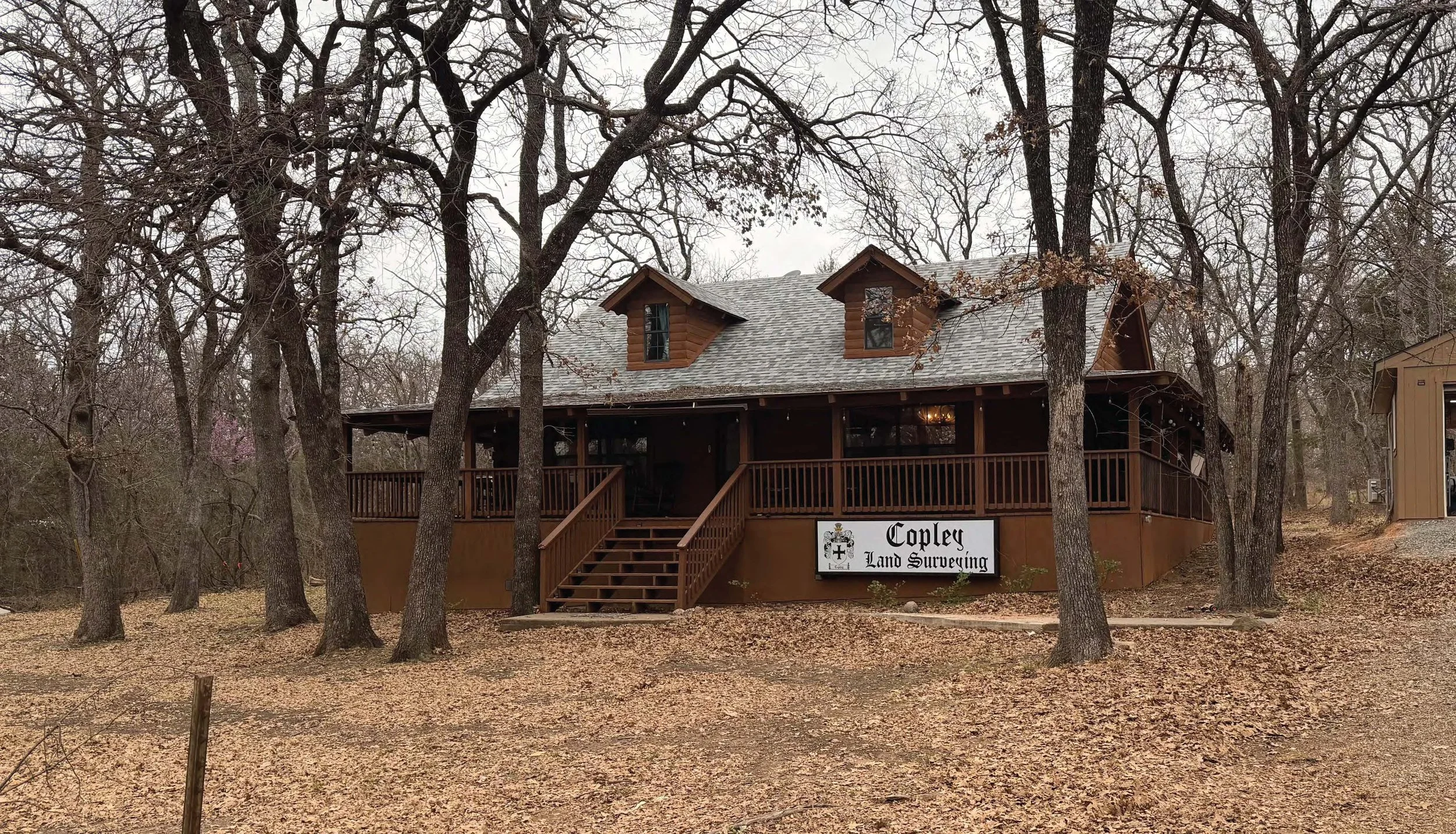 A two-story wooden house surrounded by leafless trees with a porch and stairs leading to the entrance. A sign in front reads 'Copley Land Surveying'.