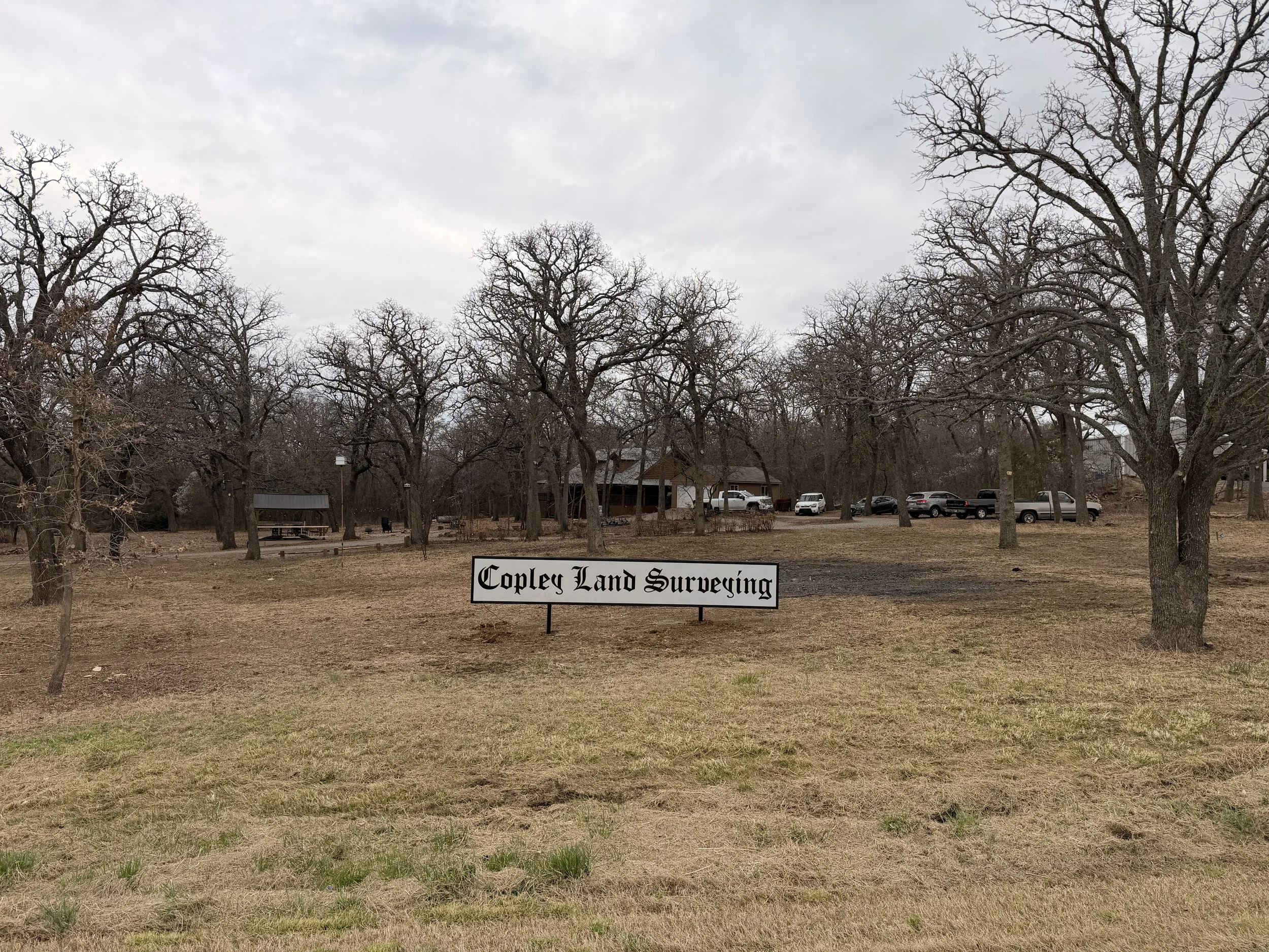 A sign in a park reads "Copley Land Surveying" with trees and parked cars in the background under a cloudy sky.