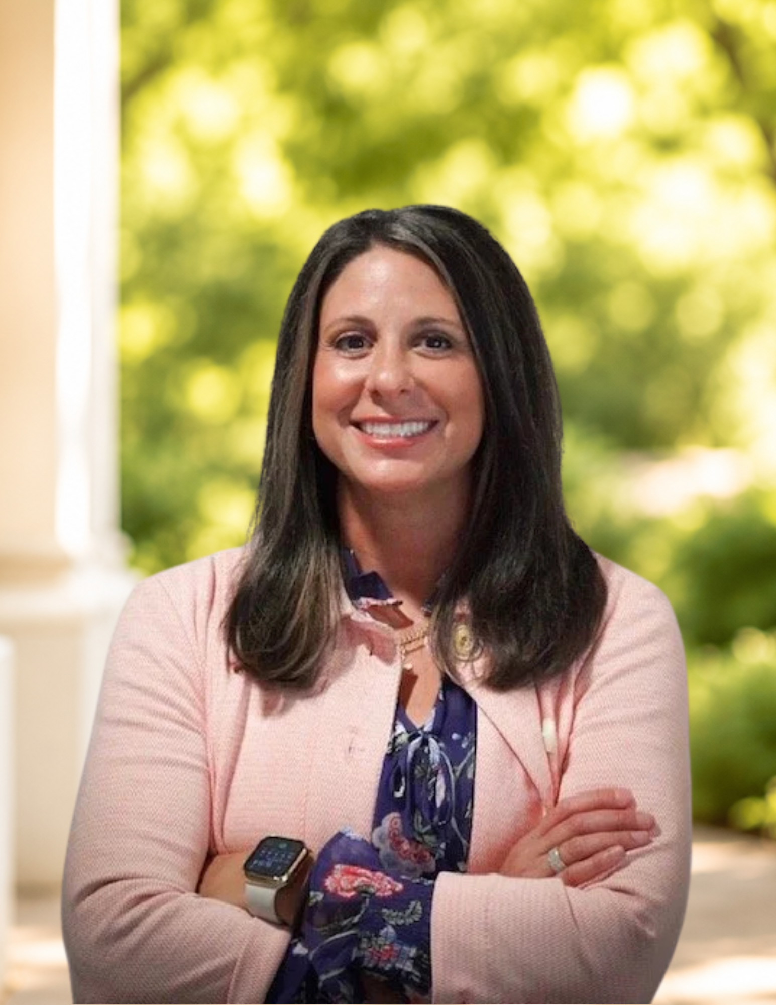 A woman smiling with arms crossed, wearing a light pink blazer, a floral blouse, and a smartwatch.