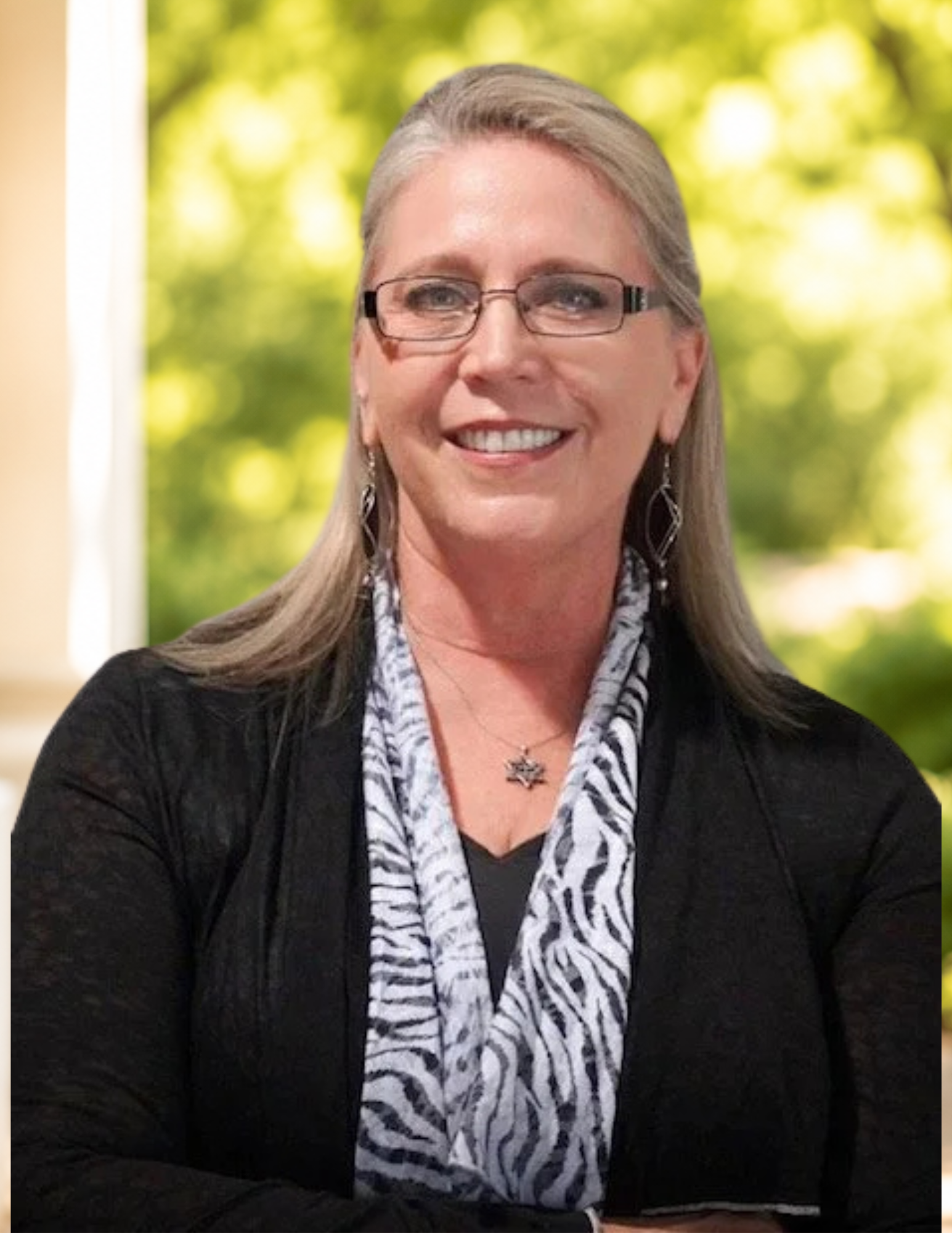 A woman with blonde hair wearing glasses, a black jacket, and a patterned scarf, smiling in front of a gray background.