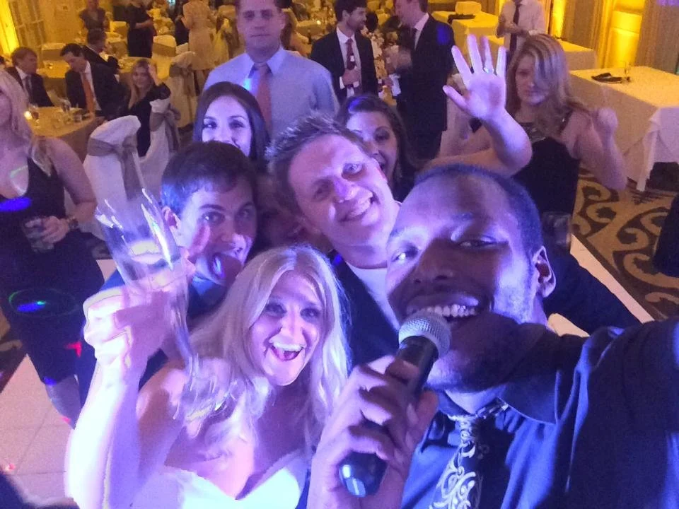 A group of people smiling and taking a selfie at a wedding reception. One person is holding a microphone, likely performing or announcing. The group is on a dance floor, with guests and tables in the background.