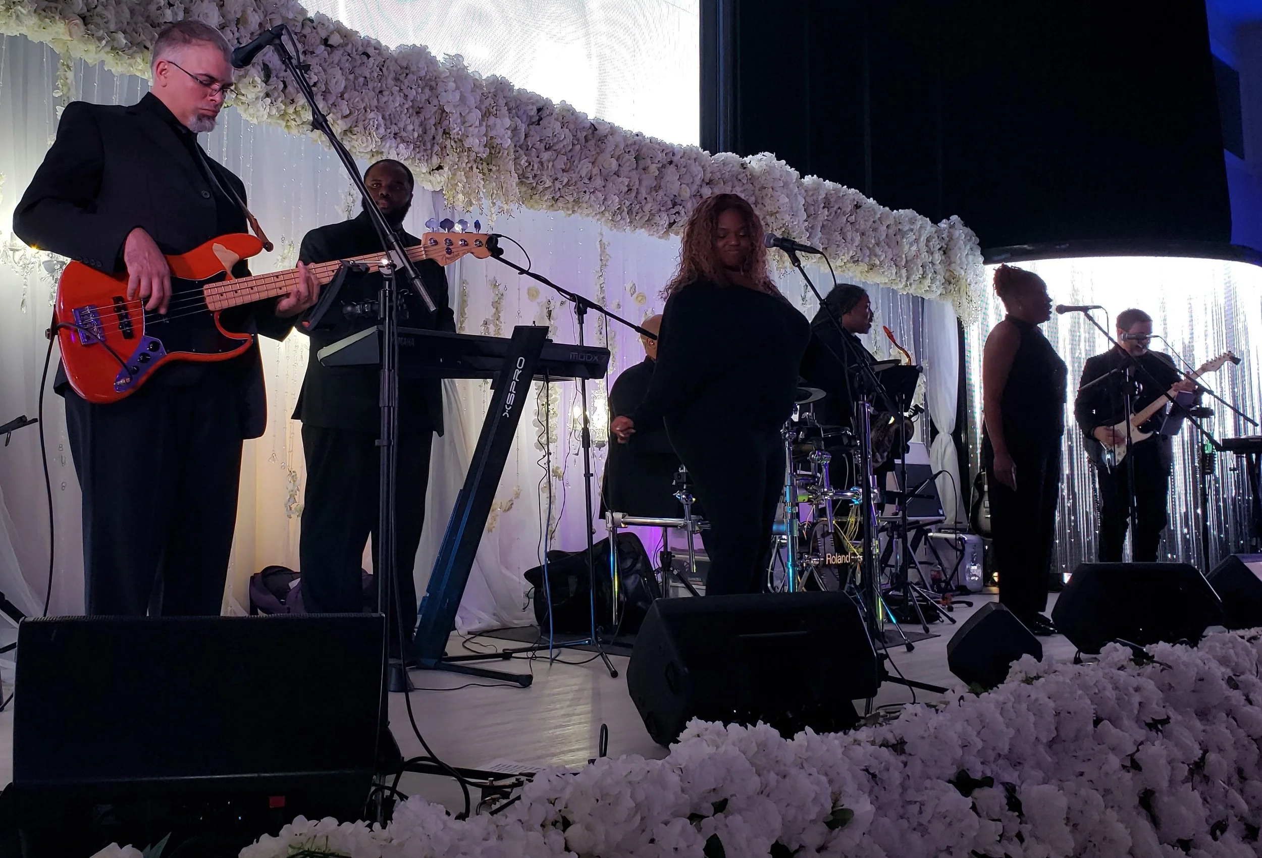 Live band performing on stage with a floral backdrop, featuring a bass guitarist, keyboardist, vocalists, and a guitarist.