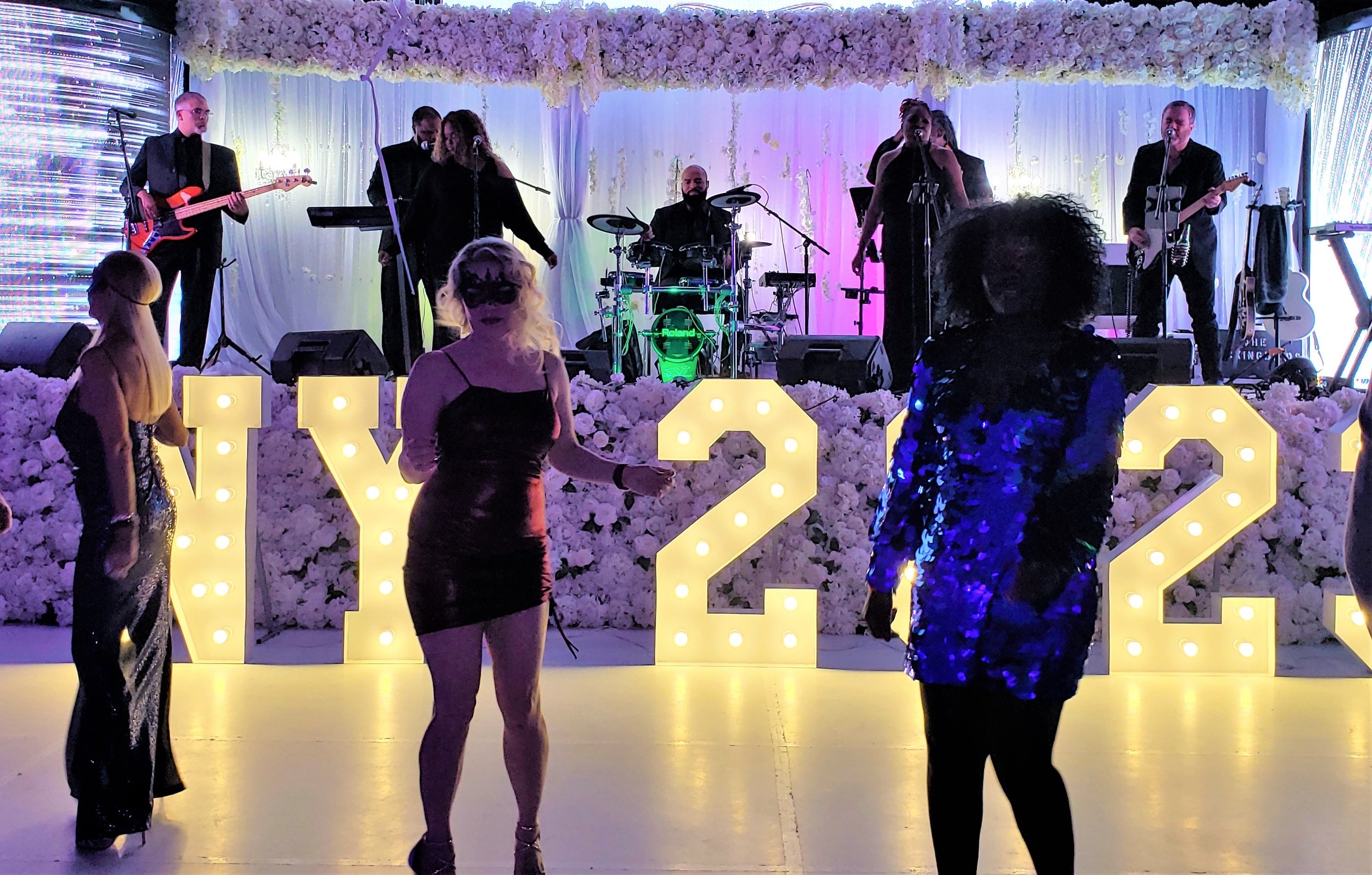 People dance at a New Year's Eve 2023 party with a live band on stage, illuminated floral decorations, and lighted numbers 'NY 2023' in the background.