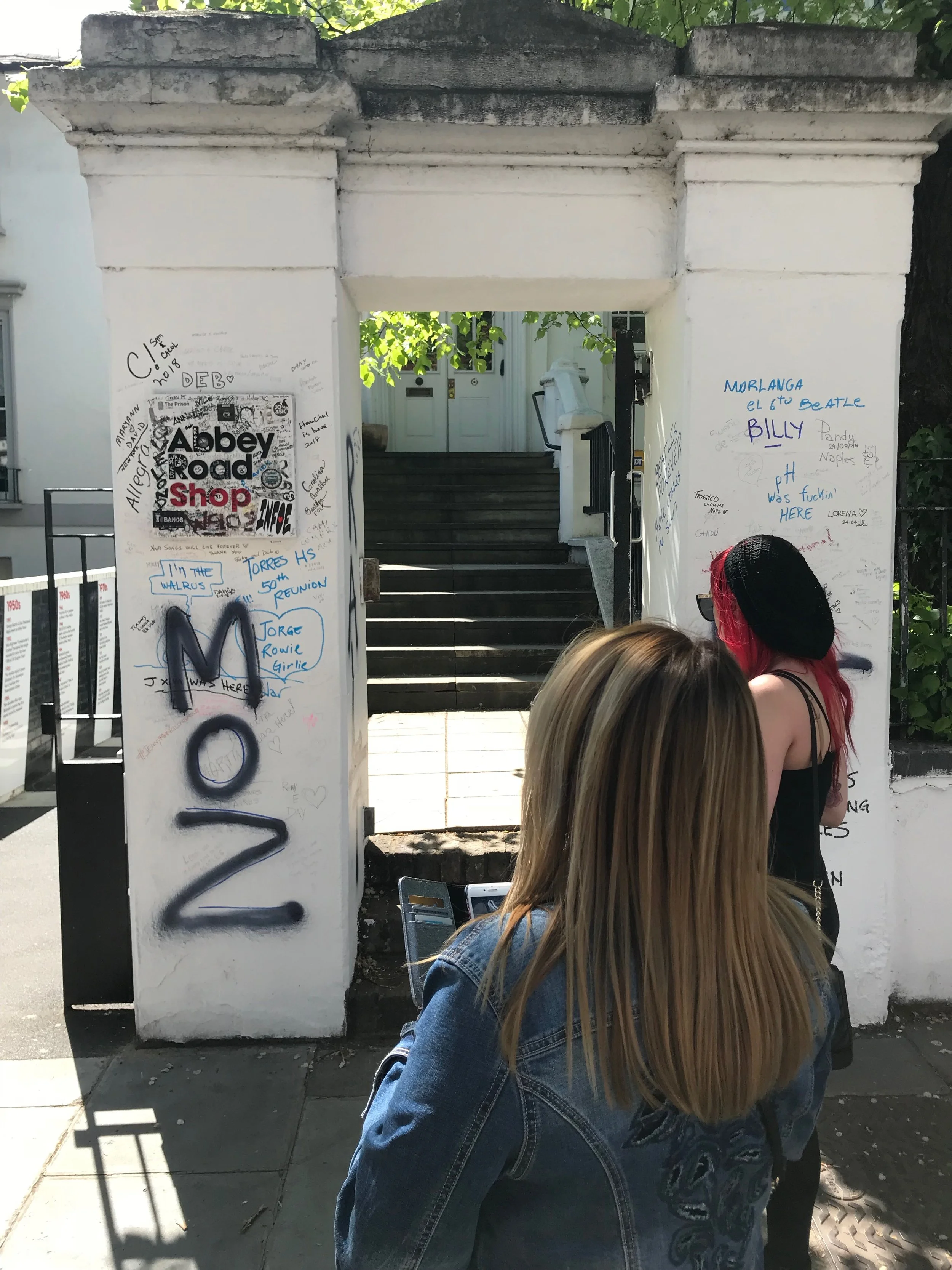 Two people stand near a graffiti-covered wall with a sign that reads 'Abbey Road Shop.' The wall is part of an entrance to a building, and various messages are written on it. Steps leading to a door are visible through the entrance.