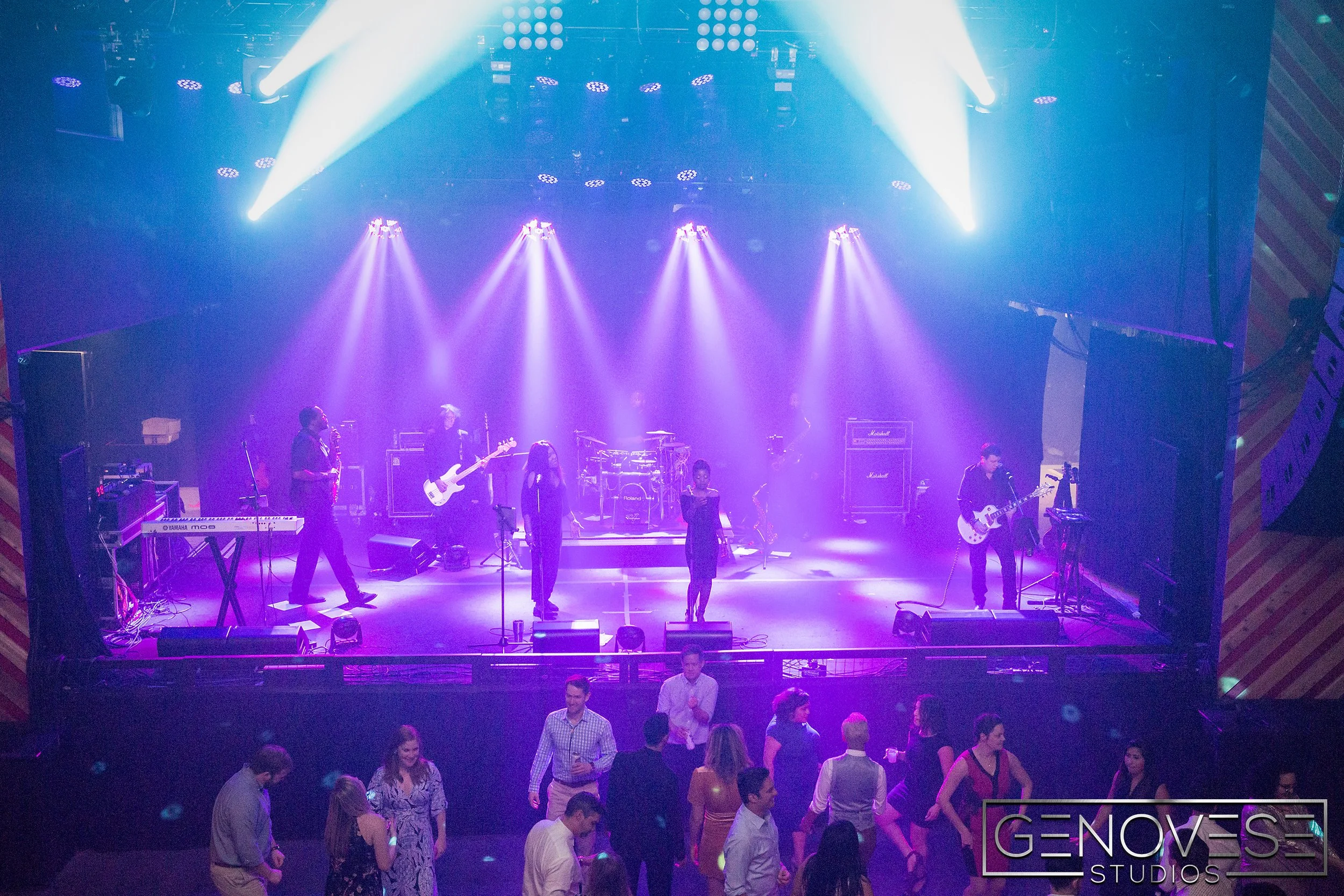 Live band performing on stage with vibrant lighting, surrounded by people dancing in front of the stage, venue has a festive atmosphere.