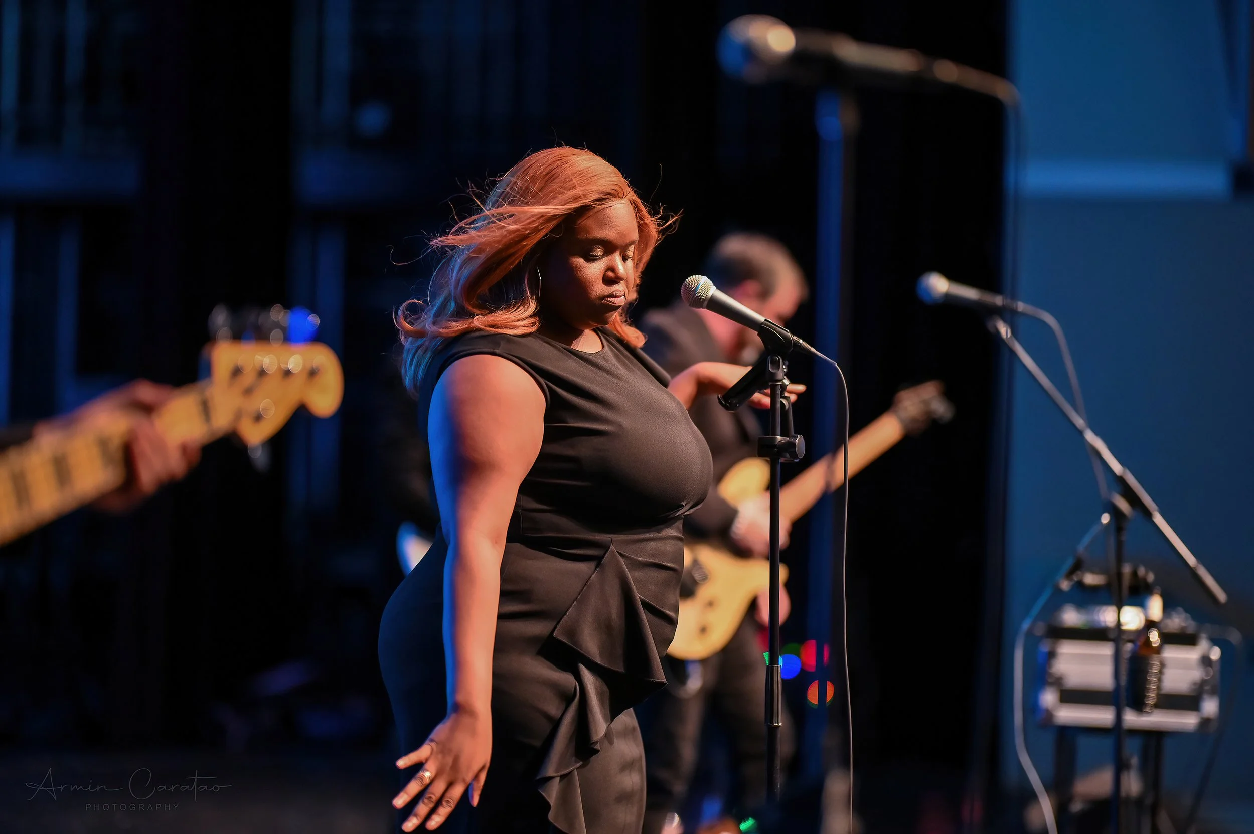 A woman singing on stage with a microphone, wearing a black dress. A musician with a guitar is partially visible in the background, along with a guitar and additional microphones.