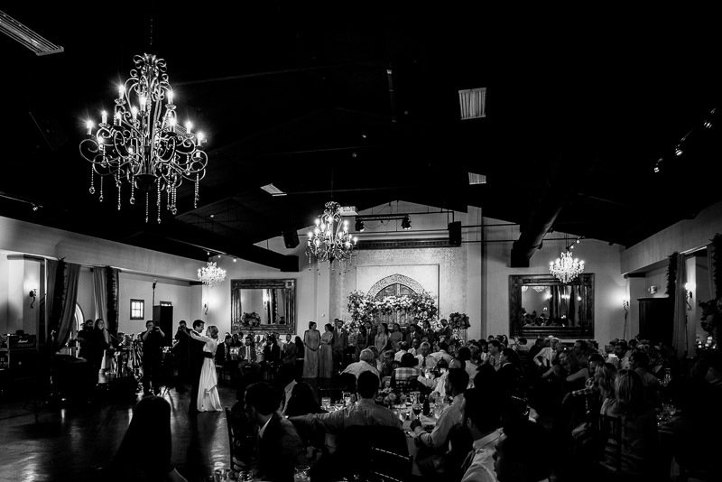 A black and white photo of a wedding reception with a couple dancing in the foreground. The room is decorated with chandeliers and floral arrangements. Guests are seated at tables observing the dance.
