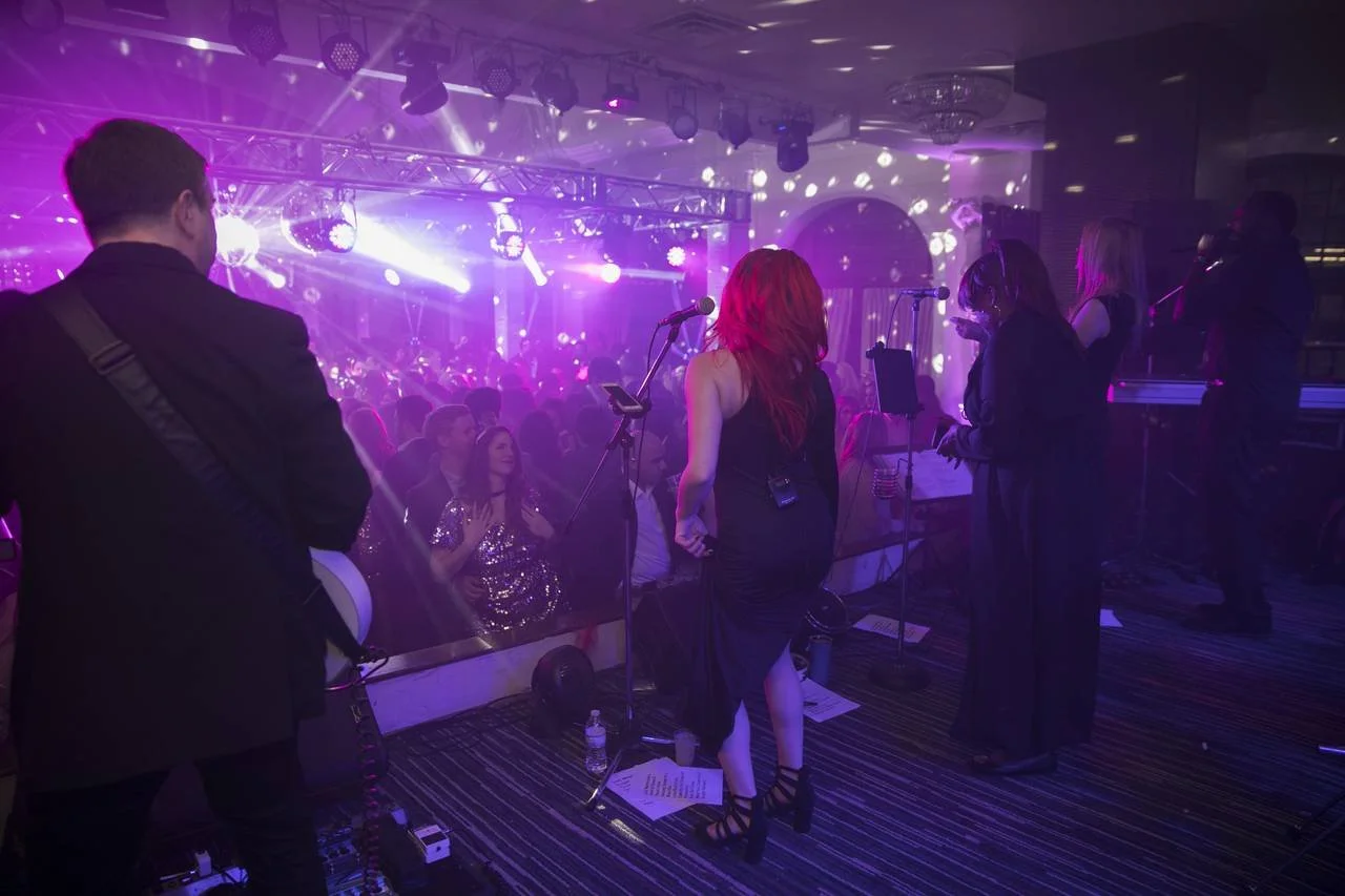 A live band performing on stage at a party with a purple lighting ambiance and a crowd of people dancing.