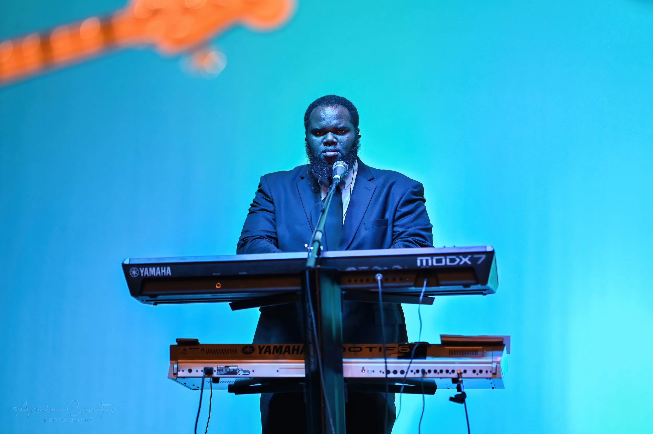 Musician playing Yamaha keyboards on stage, microphone in front, blue background.