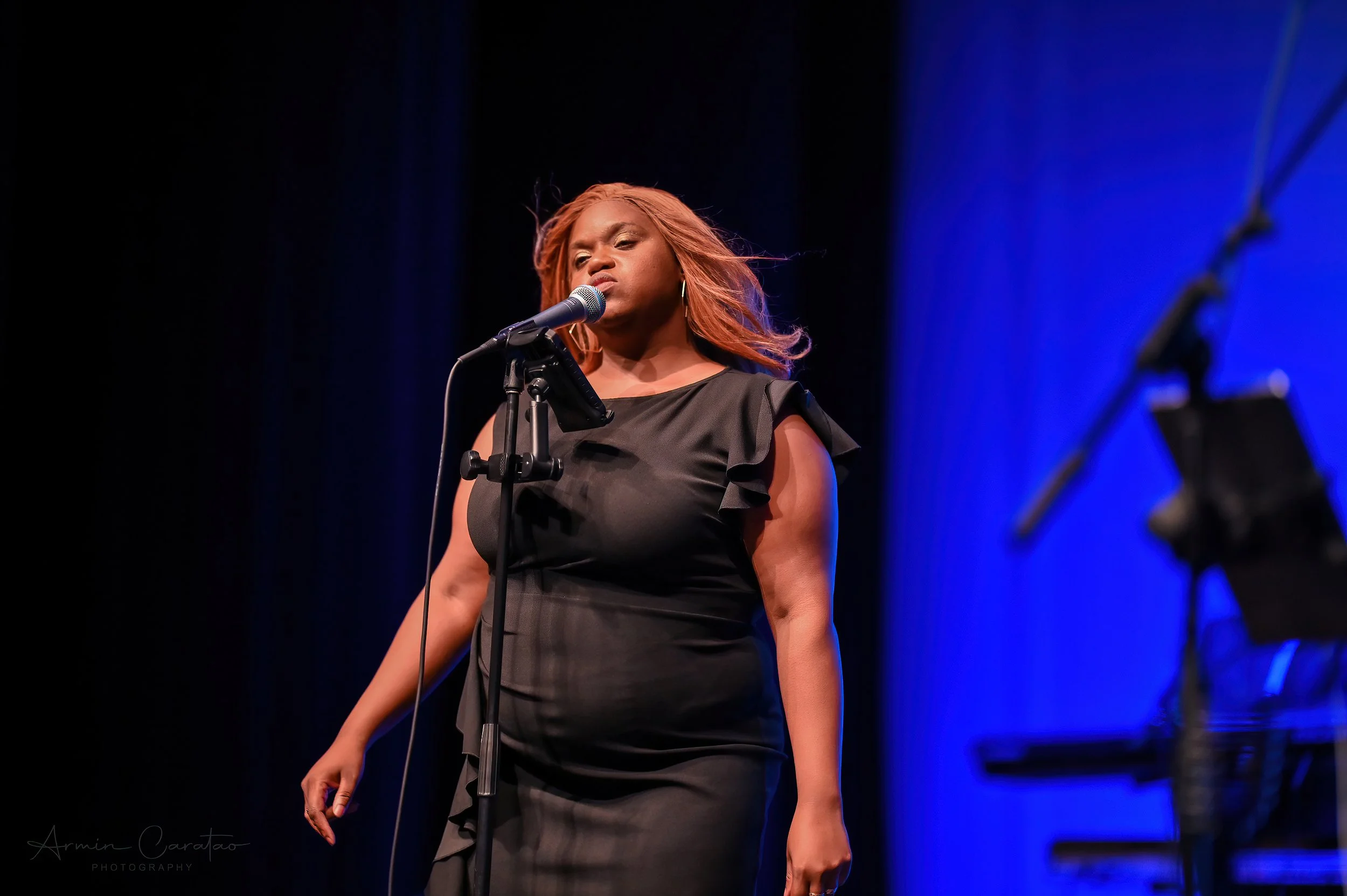 A woman in a black dress sings into a microphone on stage with a blue background.