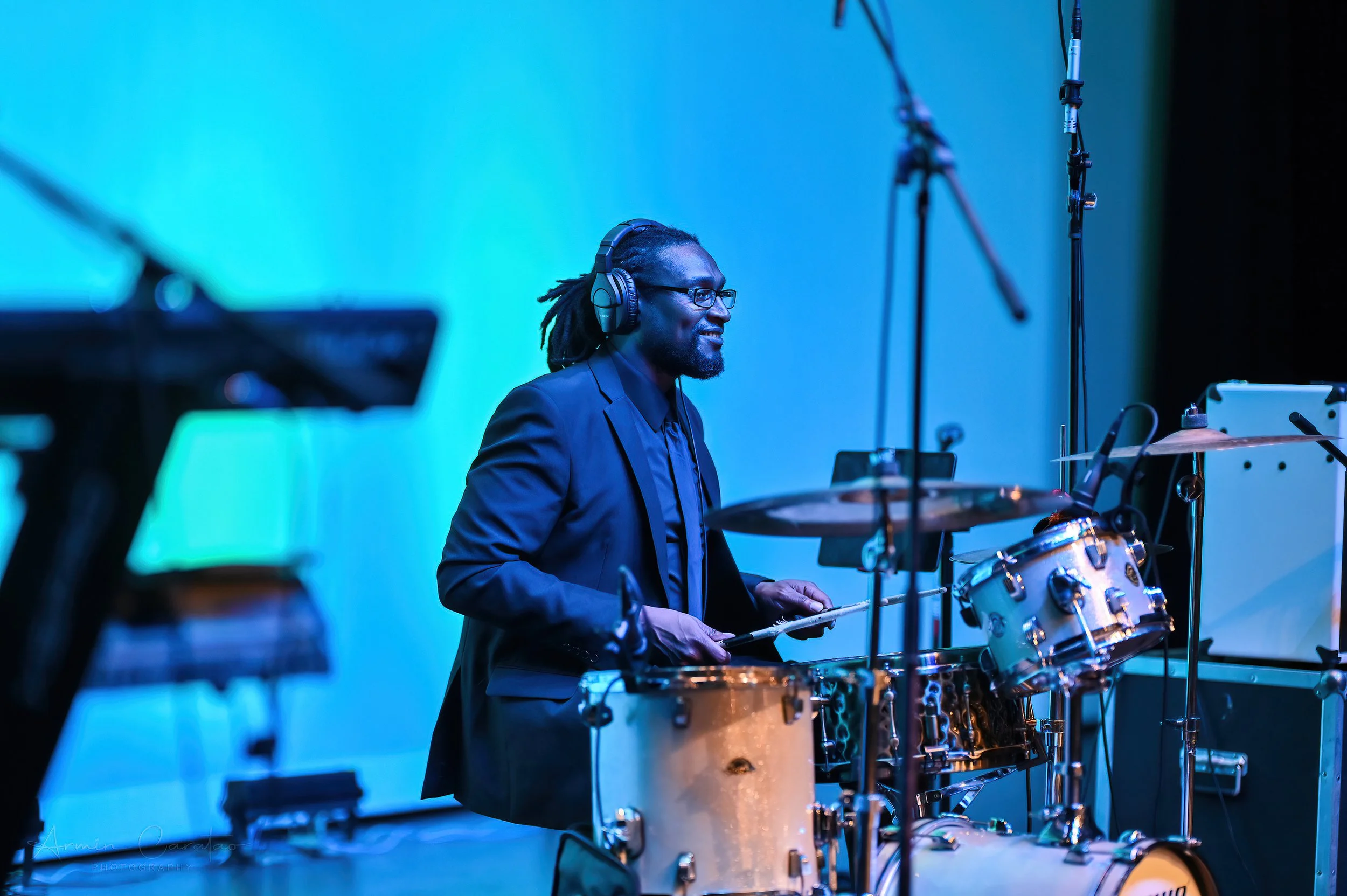 Drummer playing a drum kit on stage, wearing headphones and glasses, with a blue-lit background.