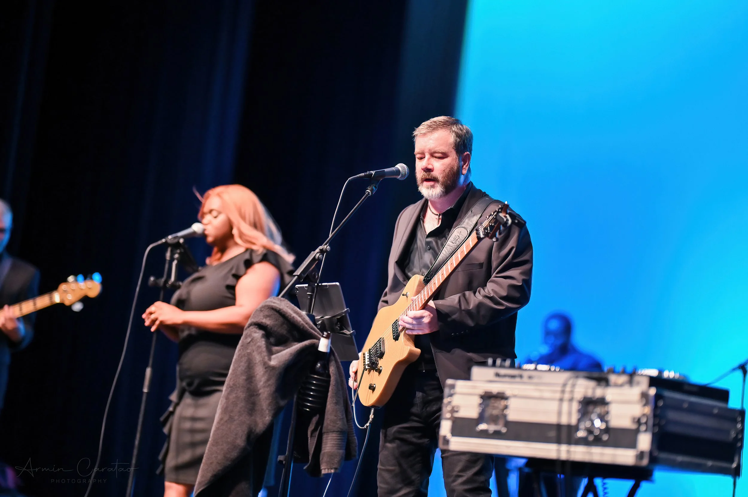 Musicians performing on stage, a woman singing into a microphone and a man playing an electric guitar, both dressed in black. A blue background is visible.