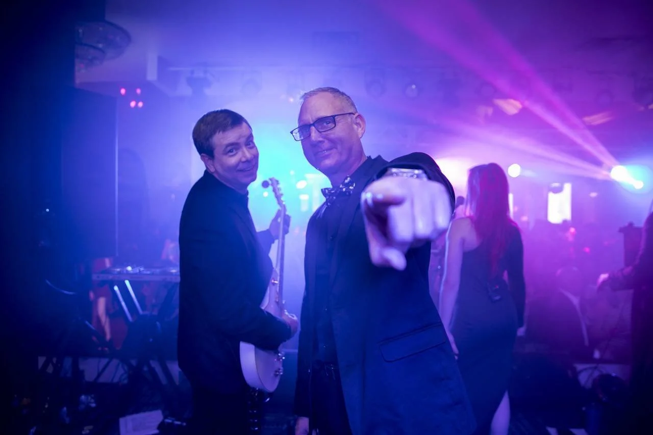 Two men in formal attire, one playing a guitar, in a club setting with colorful lights and people dancing.