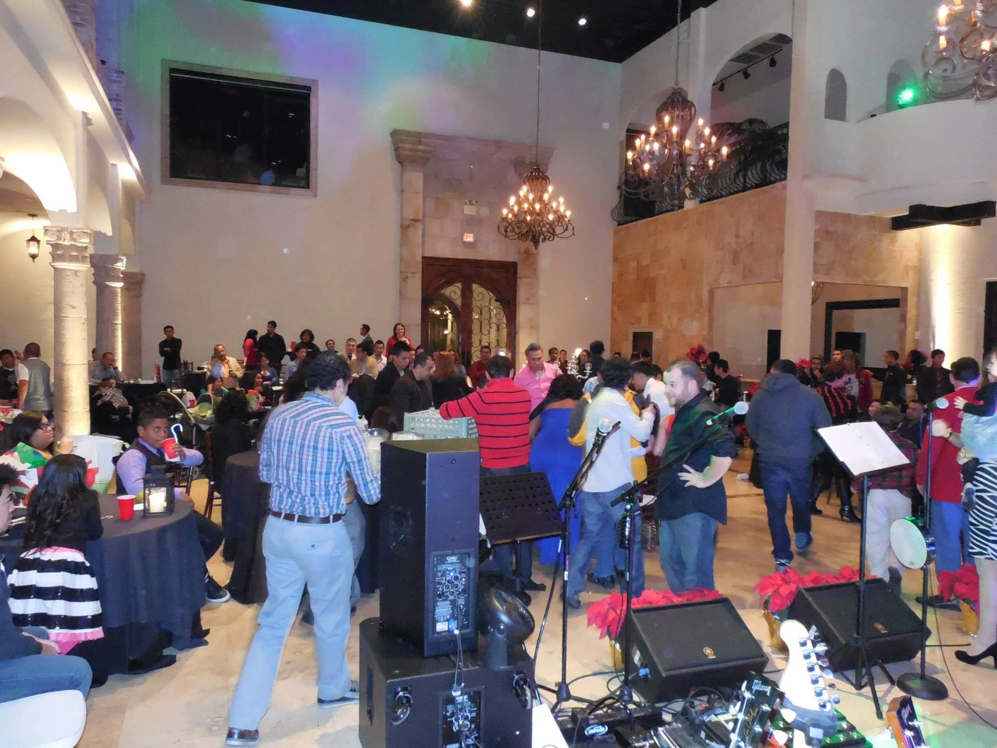 Indoor event with people socializing, tables with drinks, and musical instruments on a stage in the foreground.