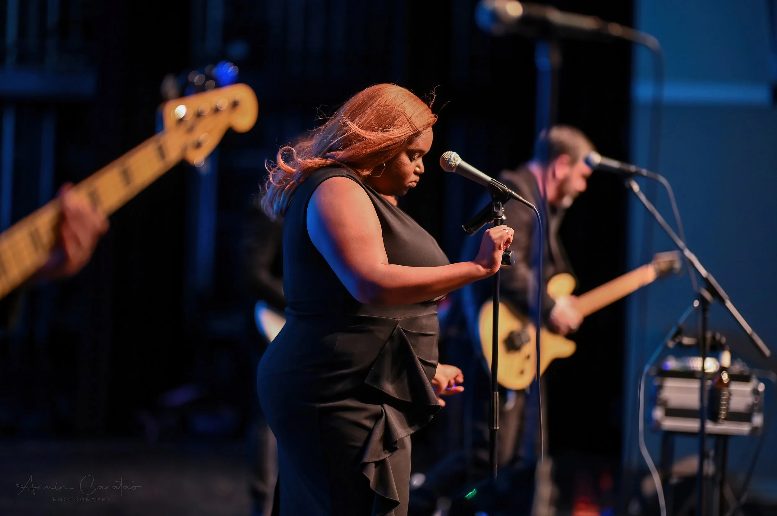 Female singer performing on stage with musicians, holding a microphone, dim lighting, wearing a black dress, guitar player in background.