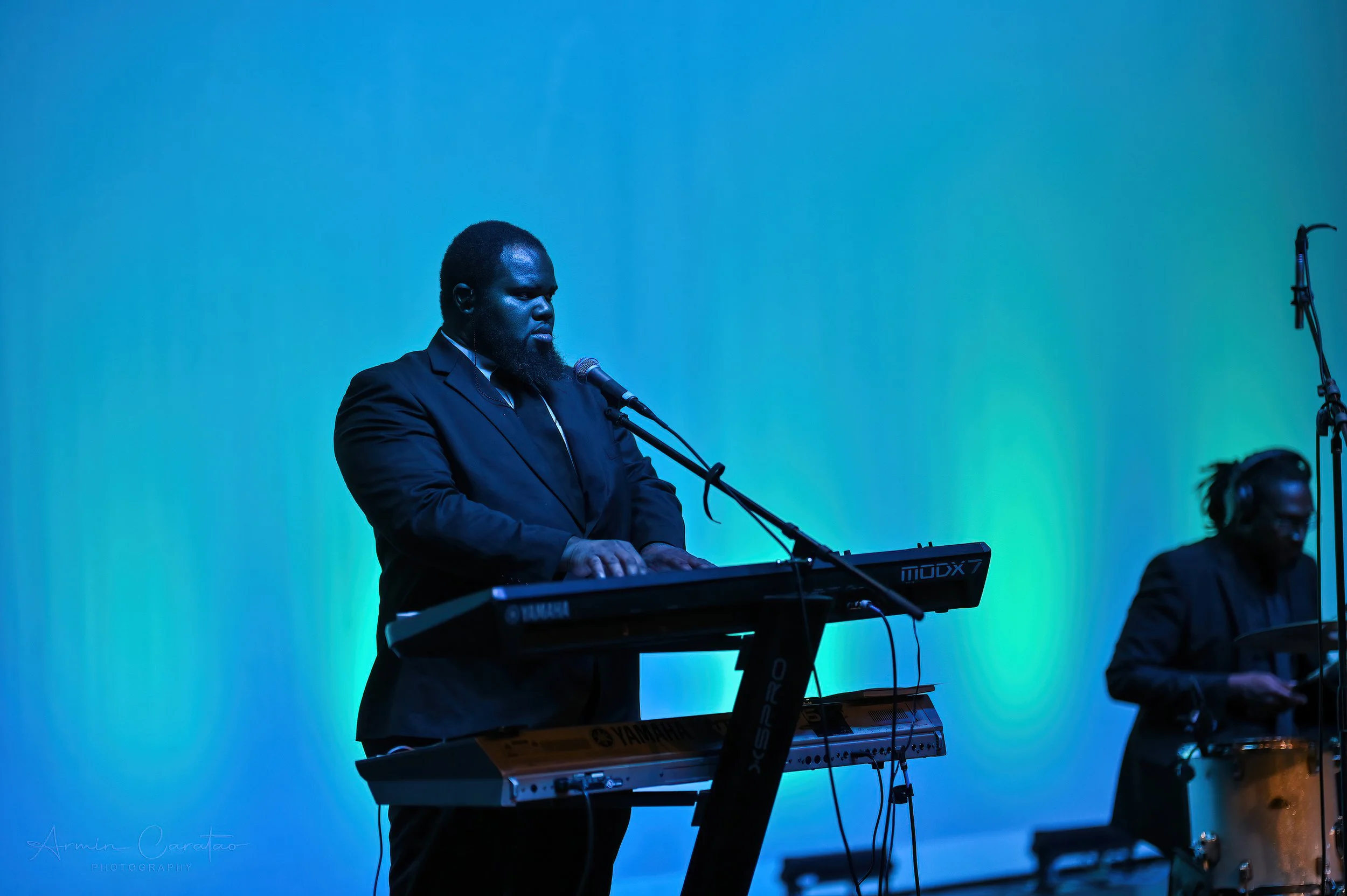 Musician playing a Yamaha keyboard and singing into a microphone on stage, with another musician playing drums in the background. Blue-green lighting.
