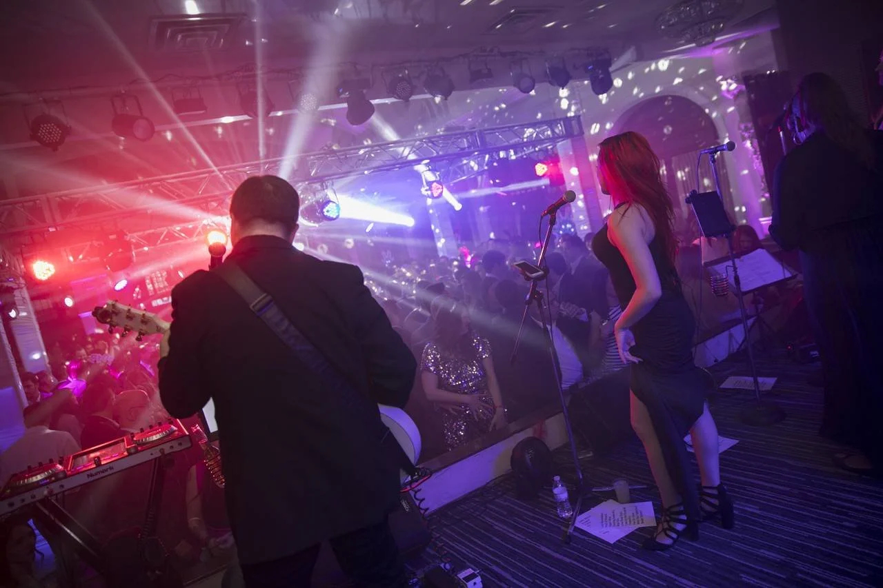 Live music performance with a band on stage, featuring a guitarist and a female vocalist with red lighting and a crowded audience in a vibrant atmosphere.