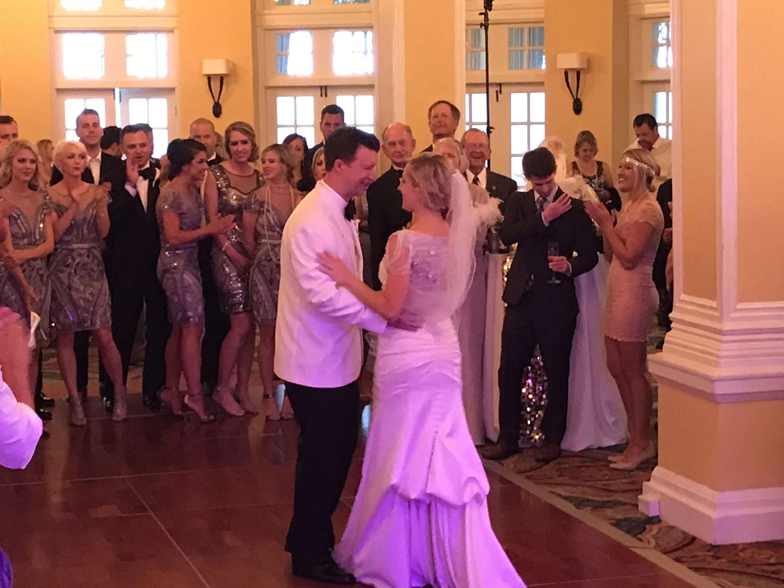 Bride and groom dancing at a wedding reception with guests watching, including bridesmaids in matching dresses and people in formal attire.