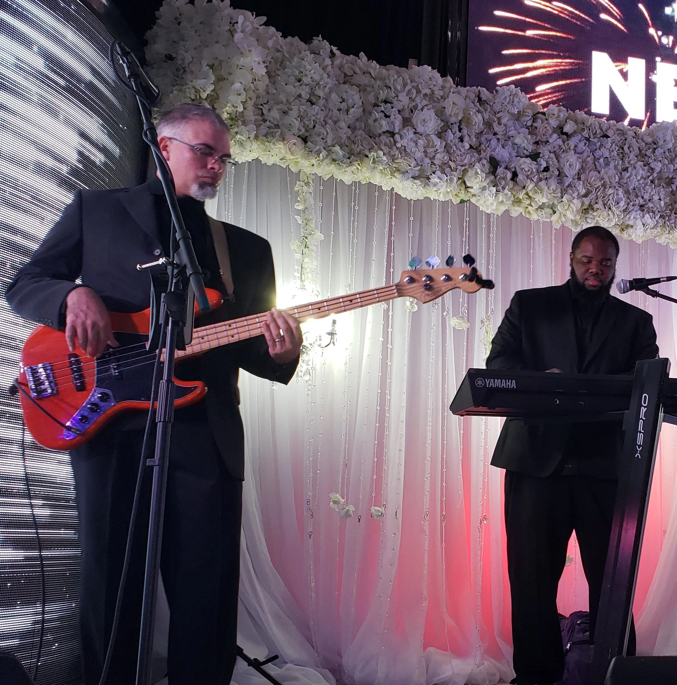 Two musicians performing on stage; one playing a red bass guitar and the other playing a Yamaha keyboard. The background features white floral decorations and a lit backdrop with text and fireworks visuals.