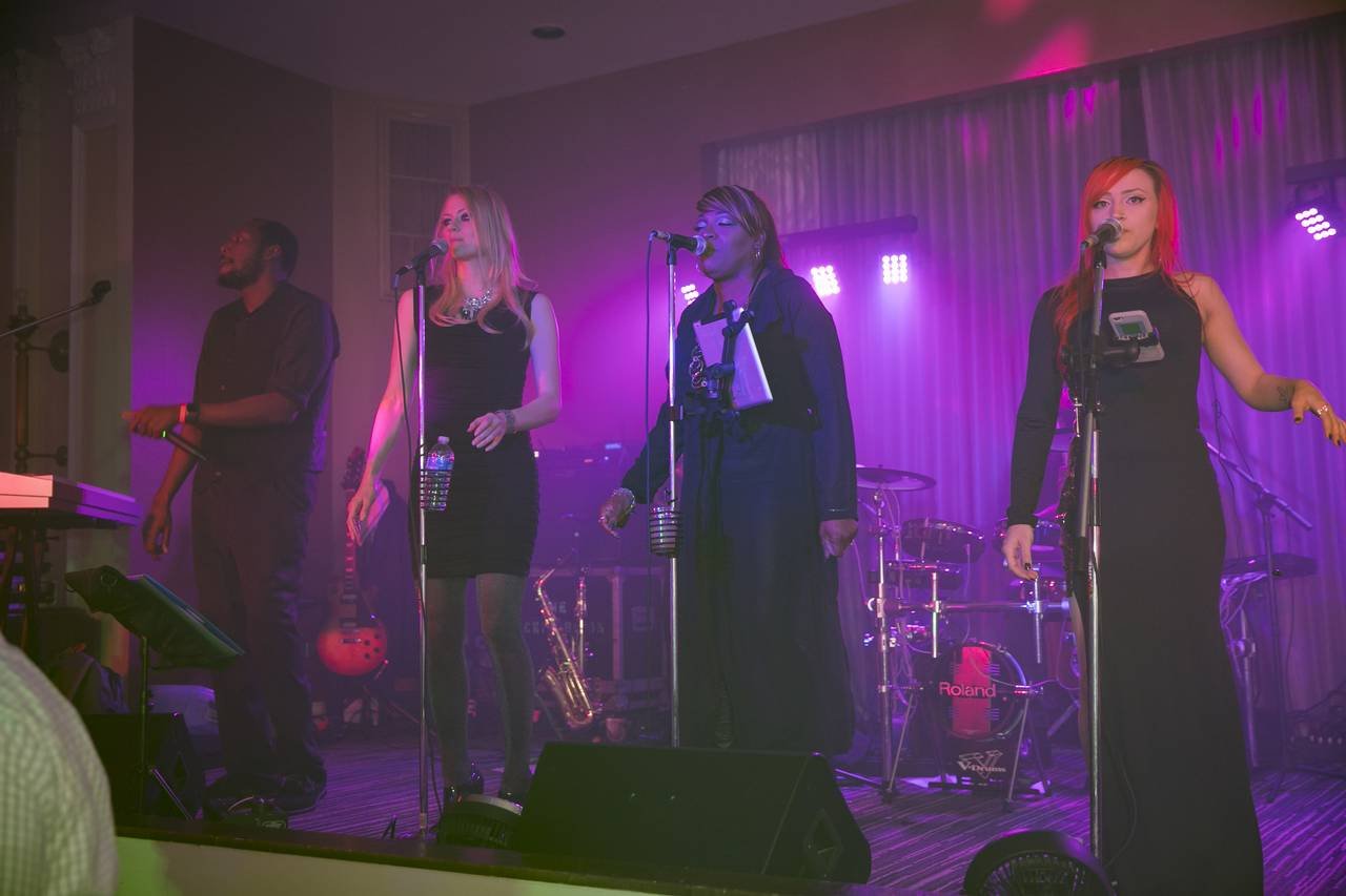 Four singers performing on stage with instruments in the background, under purple stage lighting.
