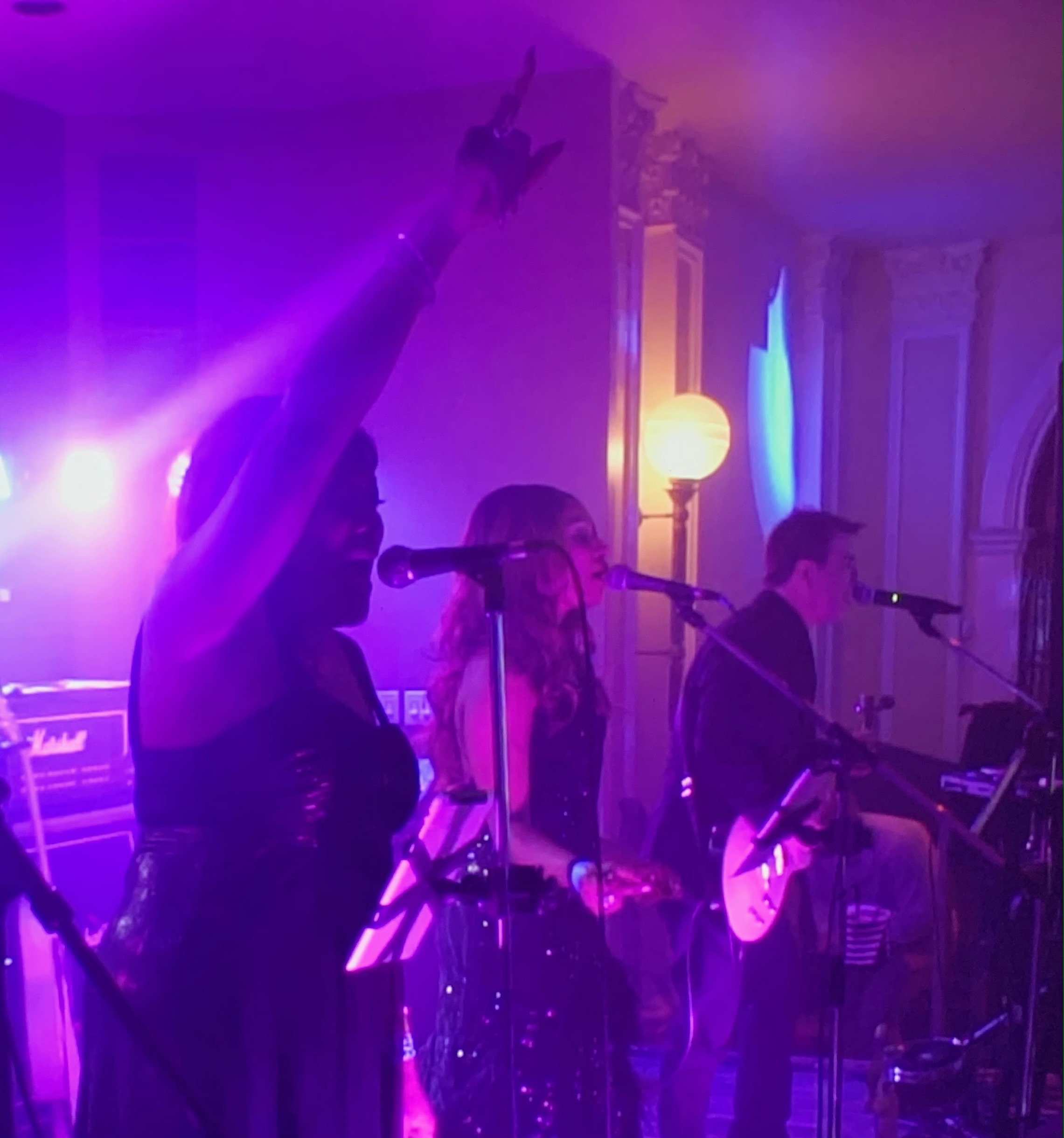 Band performing on stage with purple lighting, featuring male and female vocalists and a guitarist.