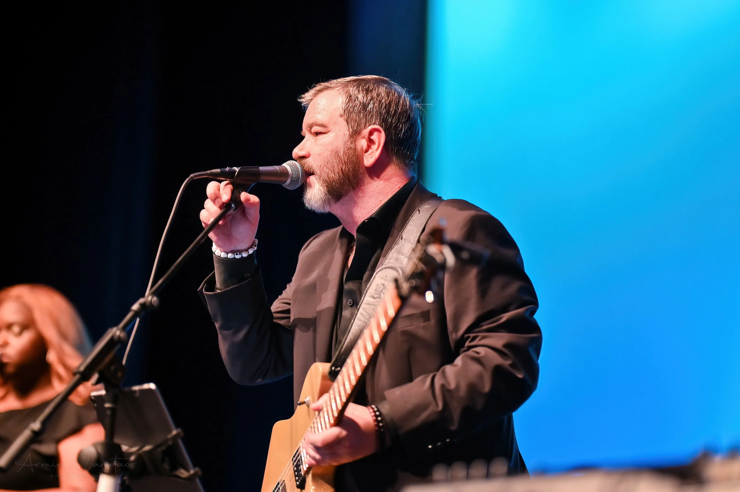 Man in a black suit singing into a microphone while playing a guitar on stage, with a female background singer blurred in the background.