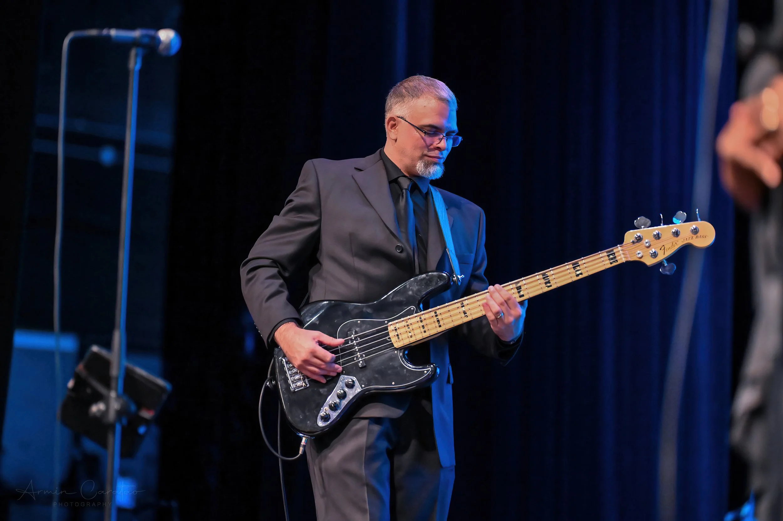 Man playing a black electric bass guitar on stage, wearing a suit, with a microphone in the foreground.