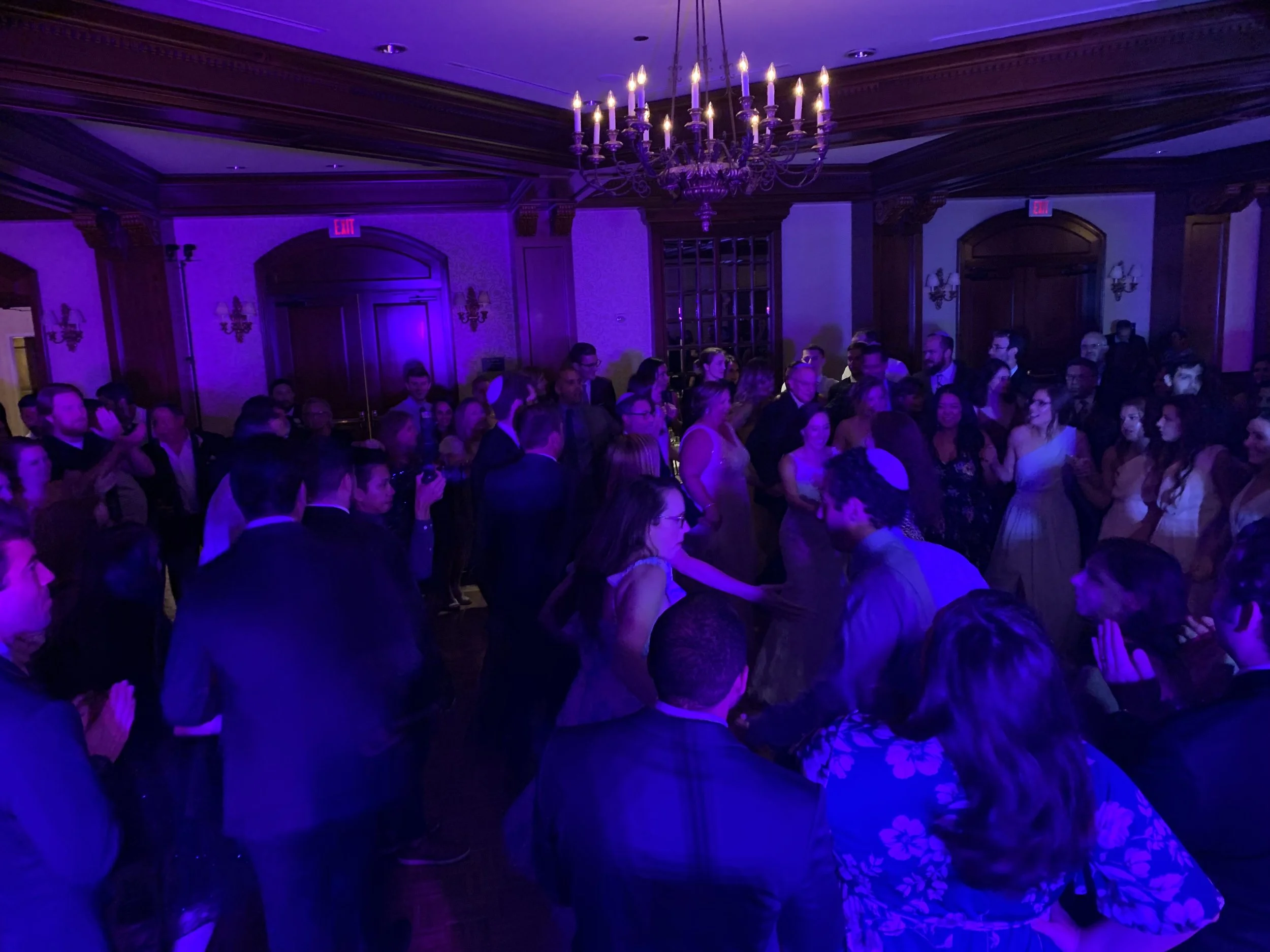 A lively indoor wedding celebration with guests dancing and mingling under purple lighting. The room features elegant chandeliers and wood-paneled walls.