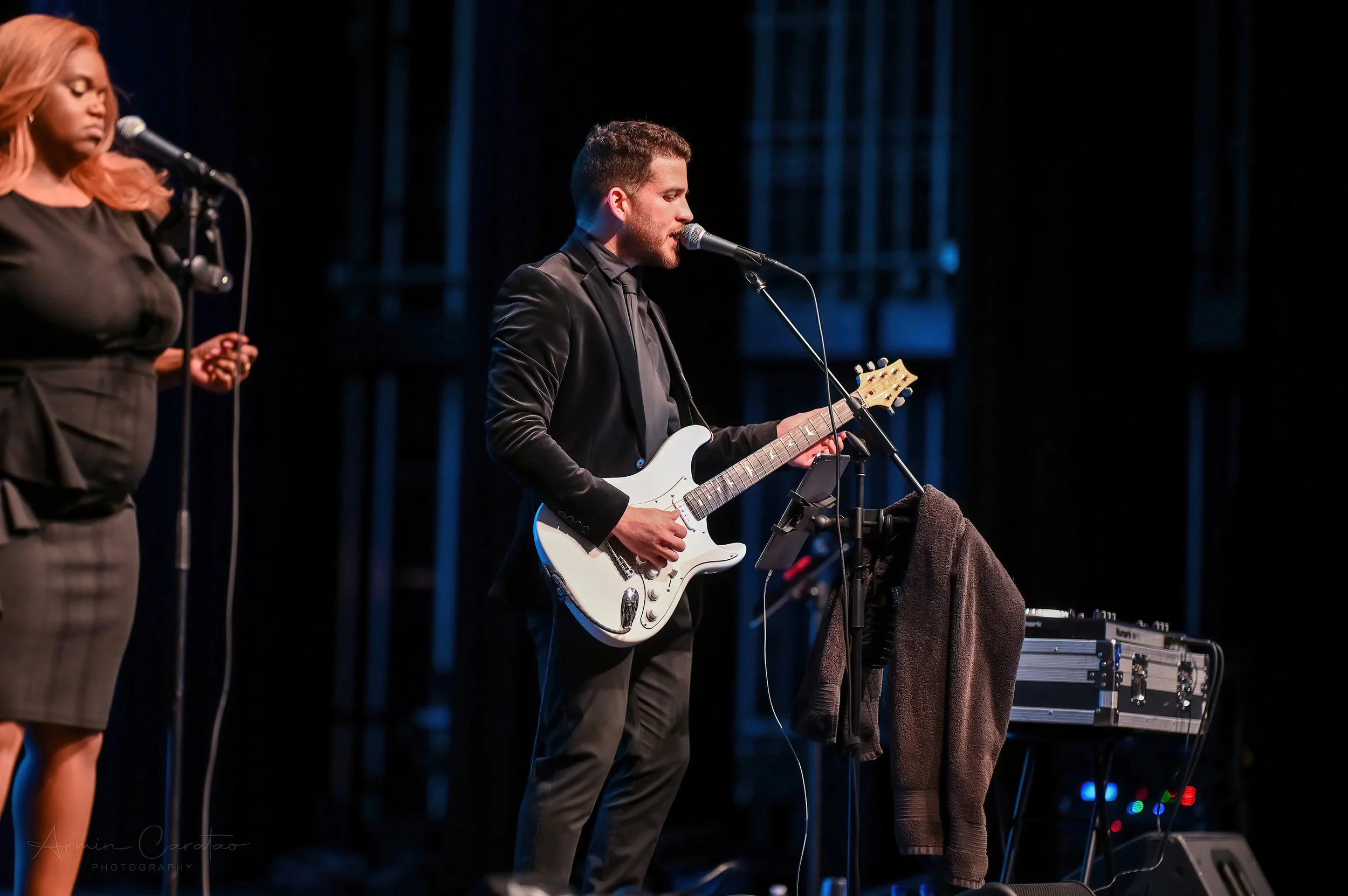 Musician playing electric guitar on stage with microphone and audio equipment, alongside a female singer.
