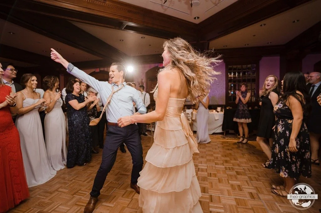 Couple dancing energetically at a wedding reception with guests clapping and smiling around them.
