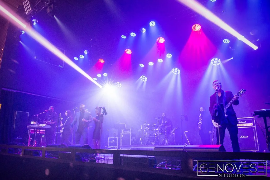 Live band performing on stage with colorful lights and musicians playing instruments, including a guitarist and saxophonist.