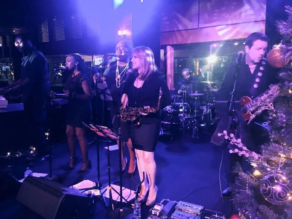 Band performing on stage with a saxophonist, guitarists, and a drummer under purple lighting.
