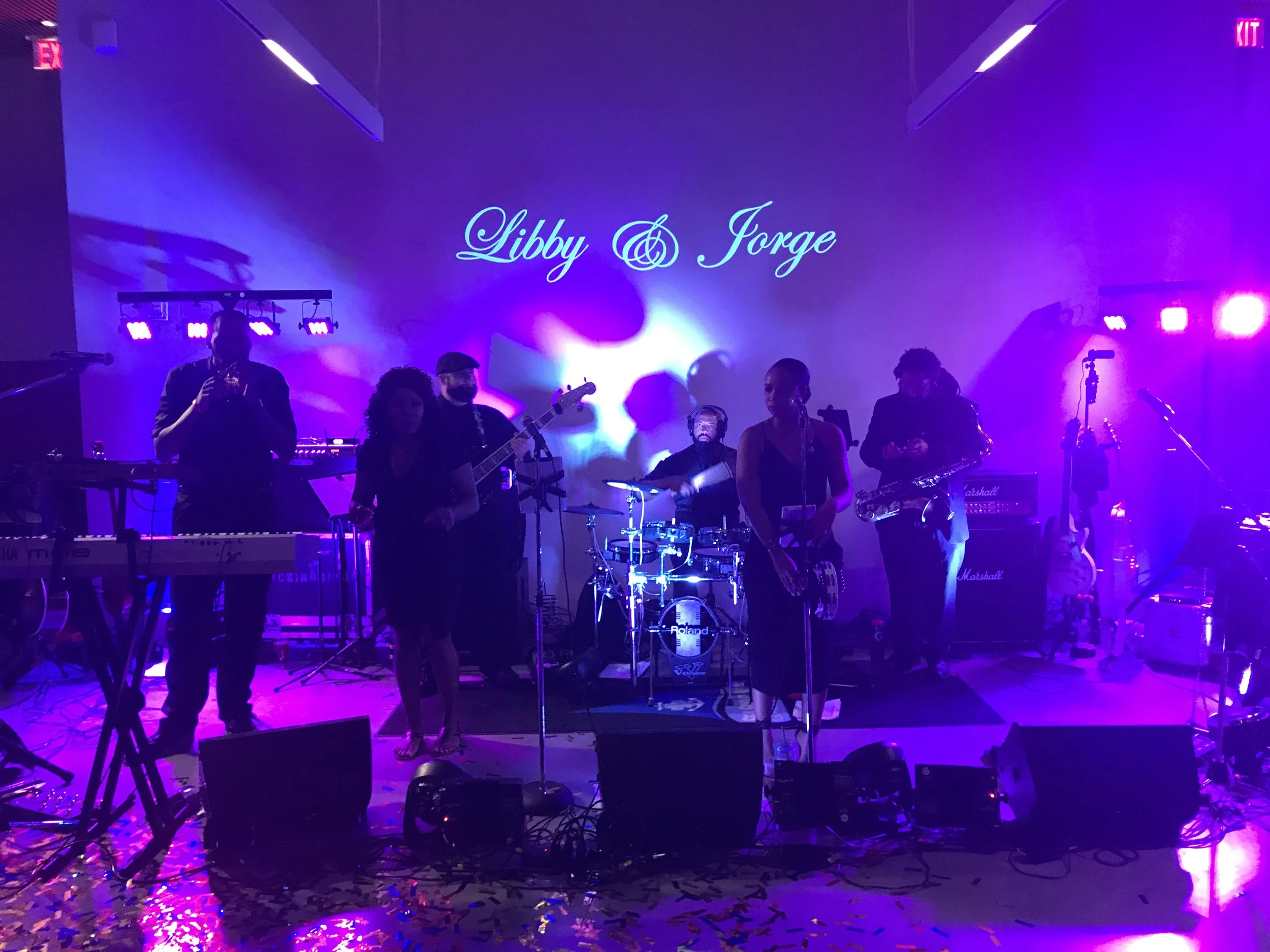 Live band performing on stage with musical instruments illuminated by purple and blue stage lights, with "Libby & Jorge" projected on the wall.