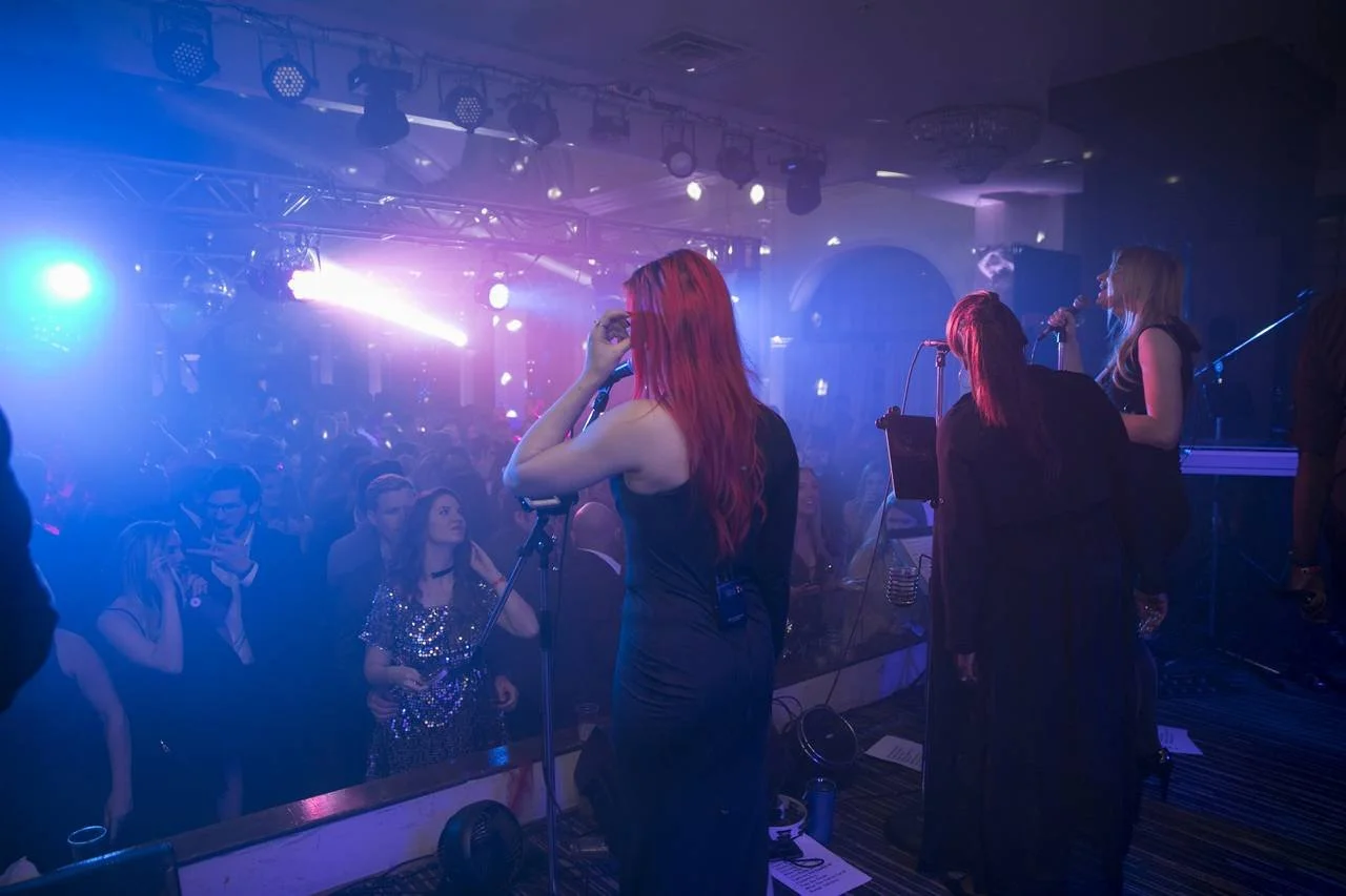 A live performance in a dimly lit venue featuring two female vocalists on stage with microphones. The room is filled with an audience enjoying the music under colorful stage lights, creating a vibrant atmosphere.