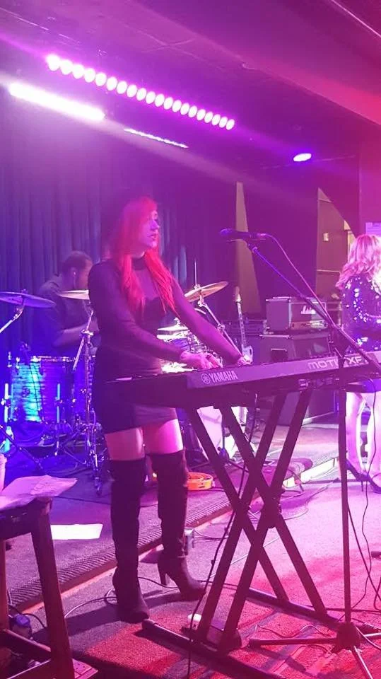 A woman playing a Yamaha keyboard on stage with a drummer in the background, under purple stage lights. She is wearing a black outfit and knee-high boots.
