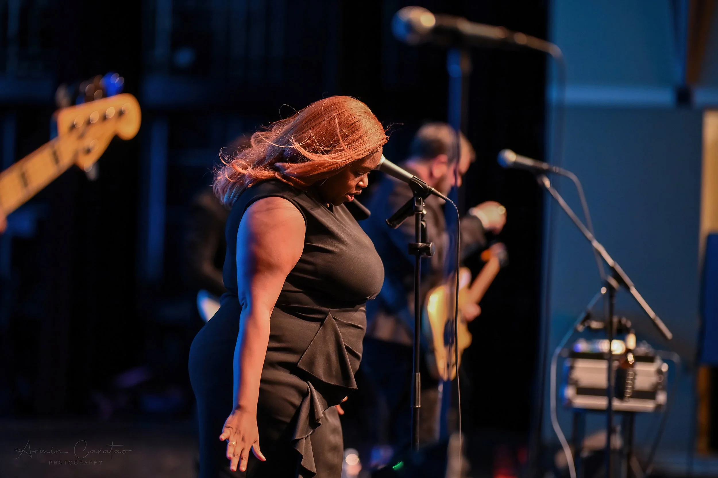 Woman singing on stage with band, holding microphone, wearing black dress; guitarist in background.