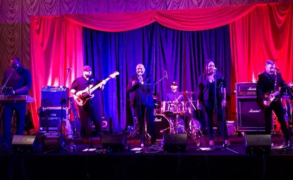 Band performing on stage with musicians playing keyboard, bass guitar, drums, and electric guitar, fronted by vocalists. Red and blue lighting enhances the backdrop.